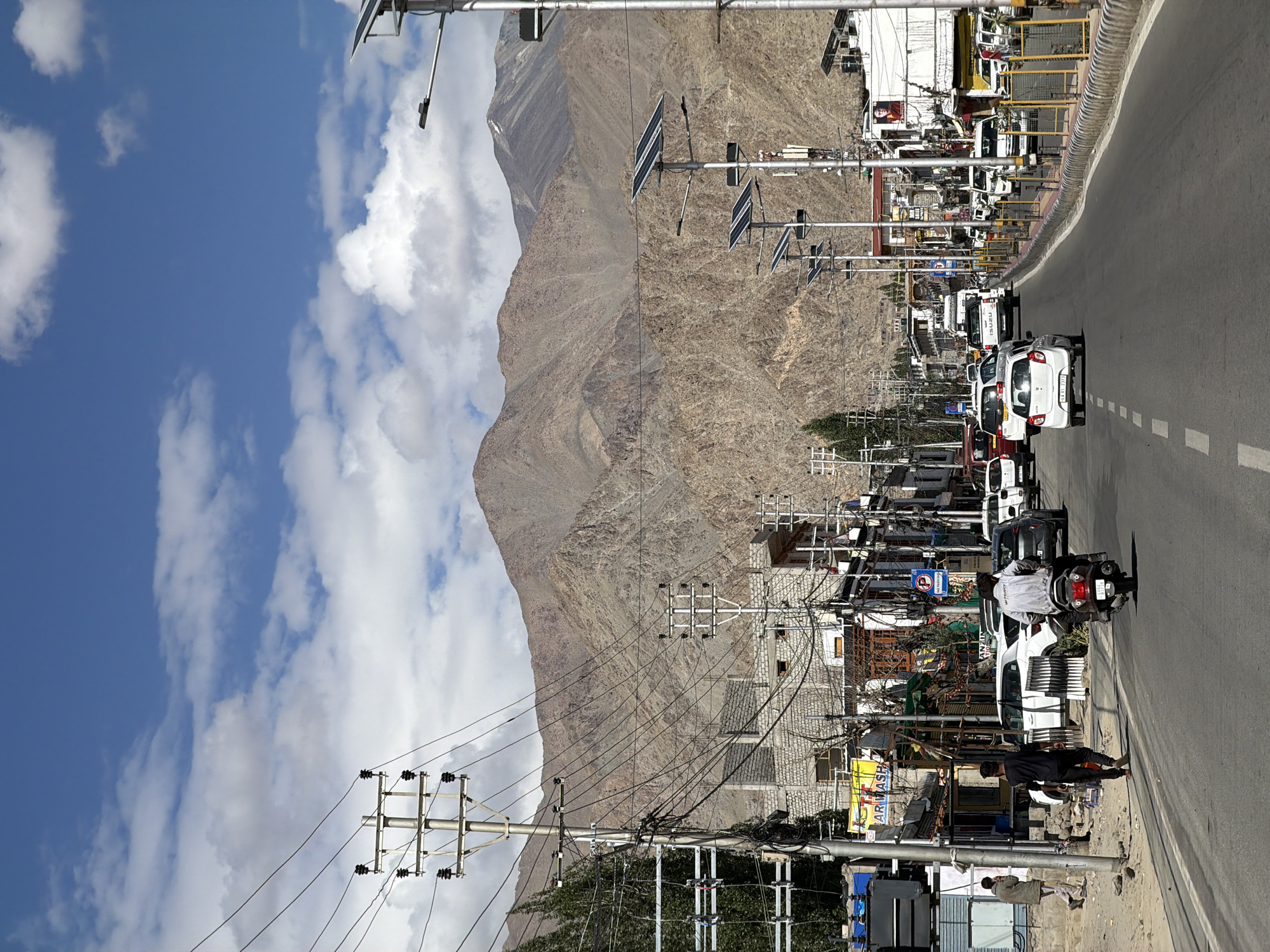 This image captures a street scene in Leh, India, with a prominent view of the Himalayan mountain range in the background. The foreground features a paved road with several vehicles, including cars and a motorcycle. On the right side of the road, there are streetlights equipped with solar panels, and a yellow barrier with reflective posts. Buildings line the left side of the road, some appearing to be shops with signage. A few individuals are visible walking on the sidewalk and riding the motorcycle. Overhead, a complex network of electrical wires and utility poles crisscrosses the scene. The sky is blue with scattered white clouds. The overall impression is that of a bustling town situated in a mountainous region.