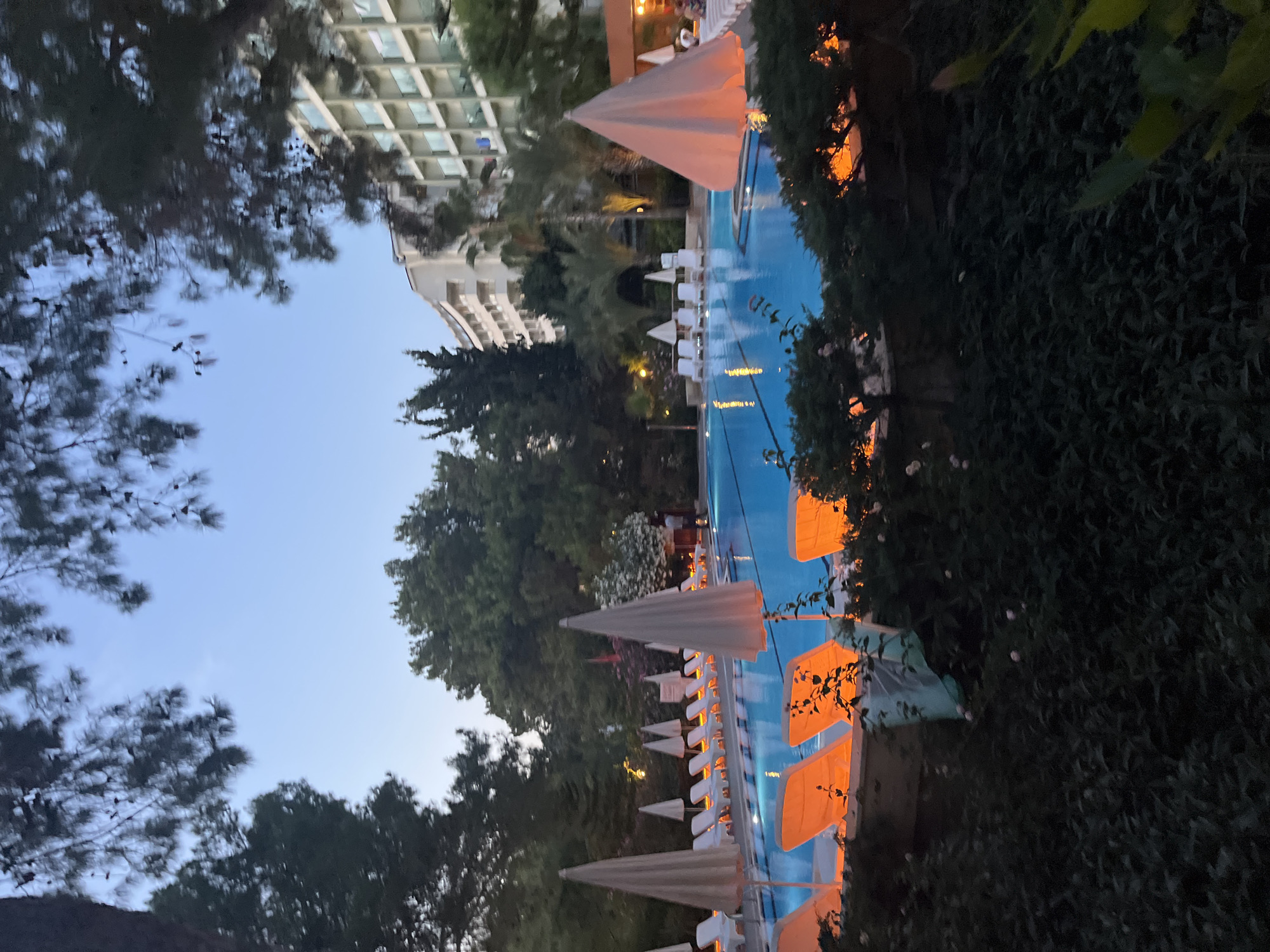 The image shows a swimming pool area at dusk, likely at a resort in Alanya, Turkey. The pool is illuminated by underwater lights, giving the water a vibrant blue hue. Around the pool are numerous lounge chairs, many of which are covered by umbrellas. Some of the lounge chairs are illuminated from beneath with an orange light, creating a warm glow. In the background, a multi-story hotel building is visible, with balconies and windows. Trees and other greenery surround the pool area, and the sky is a deep blue, indicating the transition from day to night. The overall atmosphere is serene and inviting, suggesting a place for relaxation and leisure.