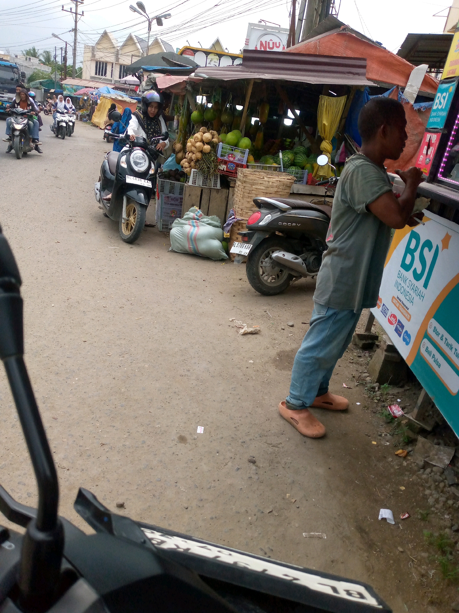 This is a street-level scene in Keudegeudong, Indonesia, likely a market or roadside area. A young man wearing a green t-shirt and blue jeans, with brown sandals, is standing by a stall with a large blue and orange banner that reads "BSI BANK SYARIAH INDONESIA". He appears to be interacting with the stall, possibly making a transaction or looking at items. In the background, there are several motorbikes, some parked and some being ridden by people wearing helmets. One motorbike in the foreground is black. The stall itself is covered with a rusty tin roof and displays various fruits like coconuts and watermelons. A large sack is leaning against a structure behind the motorbike. The ground is dirt and appears slightly damp in places. The overall impression is of a busy, open-air commercial area.