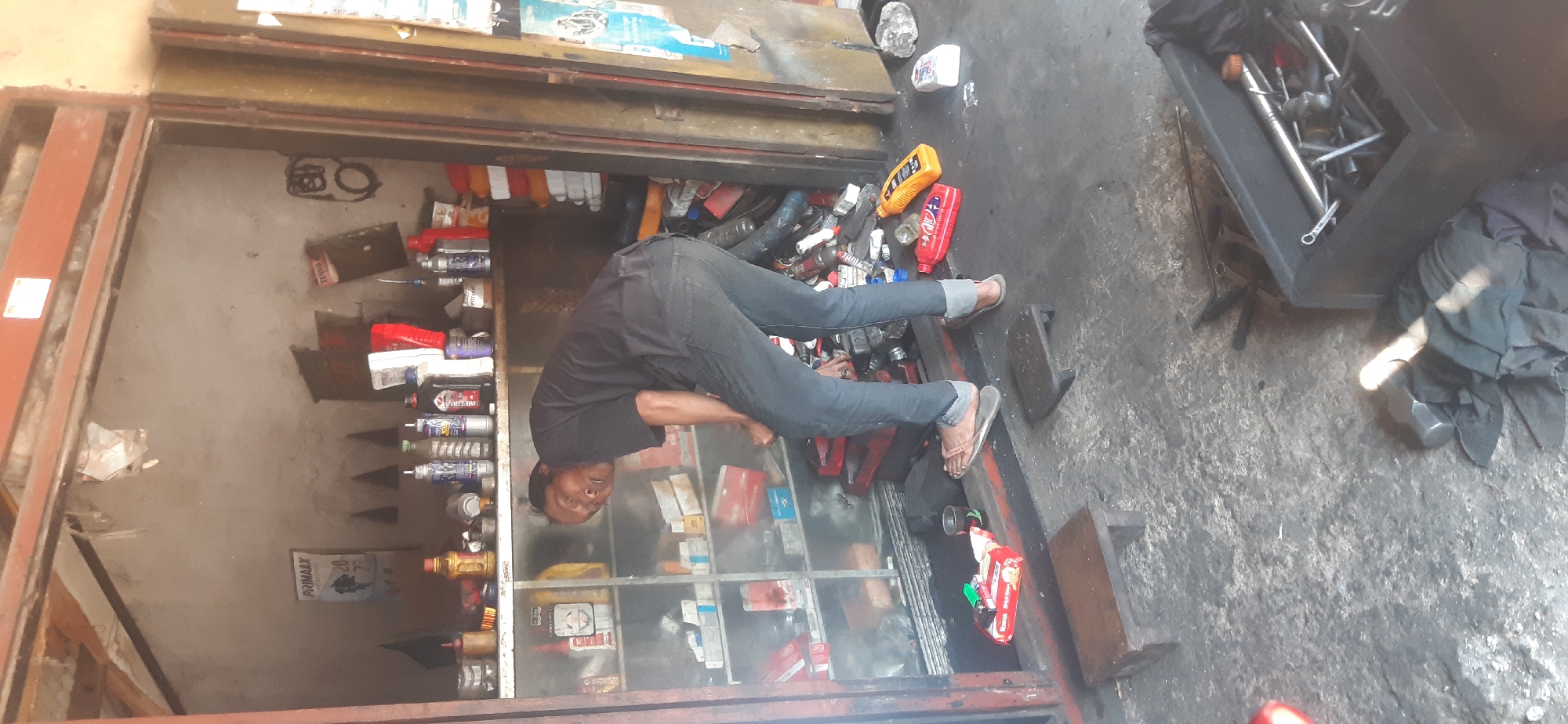 In Sepakek, Indonesia, a man wearing a black t-shirt and blue jeans is stooped over in a cluttered garage, looking at a shelf filled with various items. The shelves are packed with bottles, boxes, and other assorted objects, suggesting a repair shop or storage area. The floor is covered in dirt and debris, and scattered around are tools, oil containers, and what appear to be spare parts. Two small wooden stools are placed on the floor in front of the shelves. The overall impression is one of a busy, working environment.
