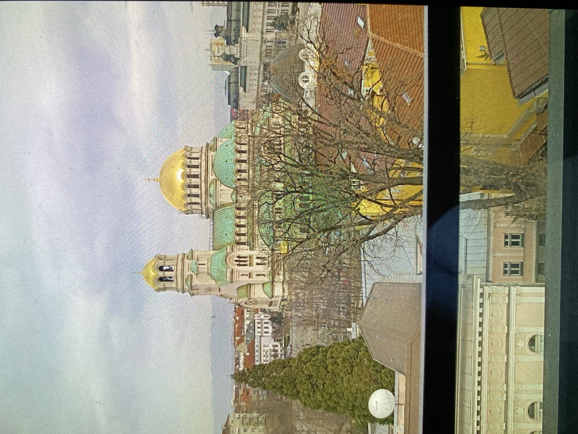 This is a daytime, panoramic photograph capturing an urban landscape, likely from an elevated perspective in Bankya, Bulgaria, possibly looking towards Sofia. The image appears to be rotated 90 degrees clockwise.
In the corrected orientation, the focal point is a magnificent, Eastern Orthodox-style cathedral positioned prominently on the left side of the frame. It features a grand, shimmering golden central dome, flanked by several smaller, distinctive verdigris-green domes. The cathedral's facade is crafted from a light-colored material, showcasing intricate architectural details such as multiple arched windows and decorative elements along its upper structure.
The foreground and mid-ground are populated with a mix of bare-branched trees, suggesting a late winter or early spring season, which partially obscure the cathedral and other buildings. A dense line of evergreen trees is visible further down towards the lower right.
The surrounding urban environment consists of various buildings with different architectural styles, some featuring traditional reddish-brown tiled roofs, others with flatter, more contemporary designs. On the far right, a bright yellow building with a dark tiled roof is visible, partially framed by a dark vertical structure, likely part of the immediate foreground, such as a window frame or balcony.
The sky is uniformly overcast and cloudy, casting a soft, diffused light across the entire scene. In the lower right corner, a white, circular satellite dish is visible. There are no people or discernible activities taking place, and no visible text is present in the image. The architectural style and prominent golden and green domes strongly suggest the cathedral is the Alexander Nevsky Cathedral, located in Sofia, Bulgaria.
