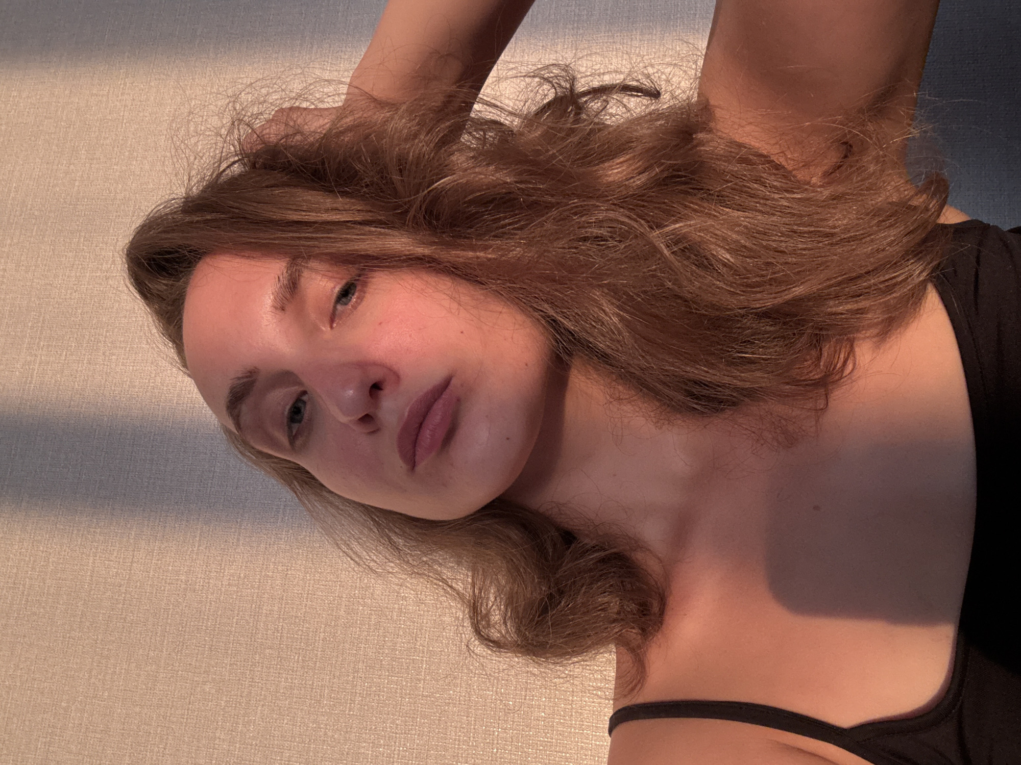 A young woman is posing for a selfie against a textured wall, with warm, directional light casting shadows across the scene. Her head is tilted, and her right hand is in her hair. She is wearing a black tank top. The background consists of a wall with a subtle pattern, and a shadow from an unseen object falls diagonally across it. The lighting suggests it is either early morning or late afternoon. The overall tone of the image is intimate and candid. There is no information in the image to suggest the location is the city of Подшивалово, Russia.