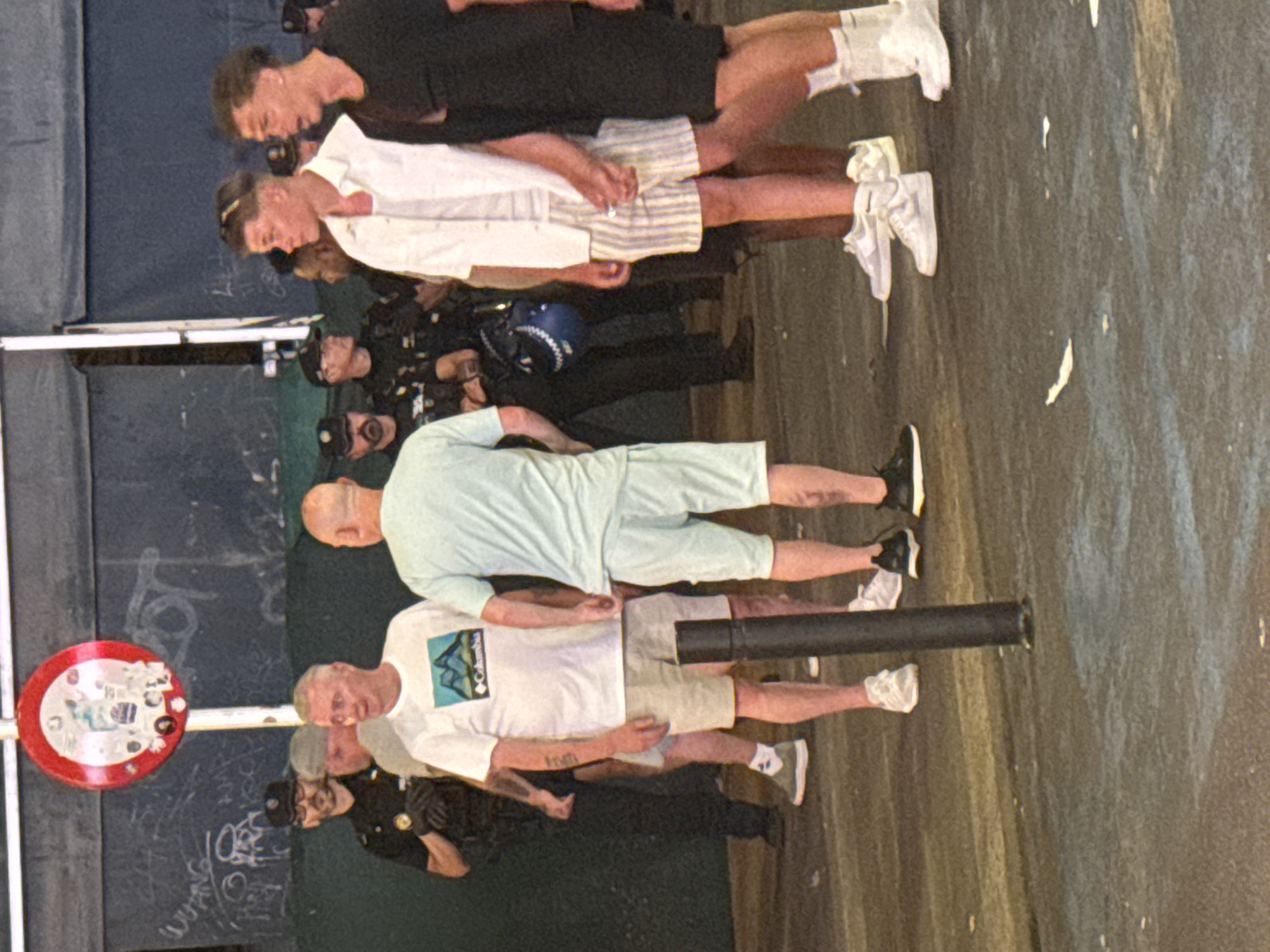 The image depicts a street scene in Benidorm, Spain. In the foreground, three men are standing. The man on the left is wearing a white t-shirt with a mountain graphic and beige shorts. The man in the center is wearing a light blue t-shirt and light blue shorts. The man on the right is wearing a white open shirt over a white undershirt and striped shorts. Behind them, several individuals in police uniforms are visible, suggesting a law enforcement presence. One police officer on the left is wearing a black uniform with a cap. A red and white circular sign with some graphics is mounted on a pole in the background. The ground appears to be paved and wet in some areas, with some white paint splatters. The overall setting suggests an outdoor public space, possibly a street or promenade.