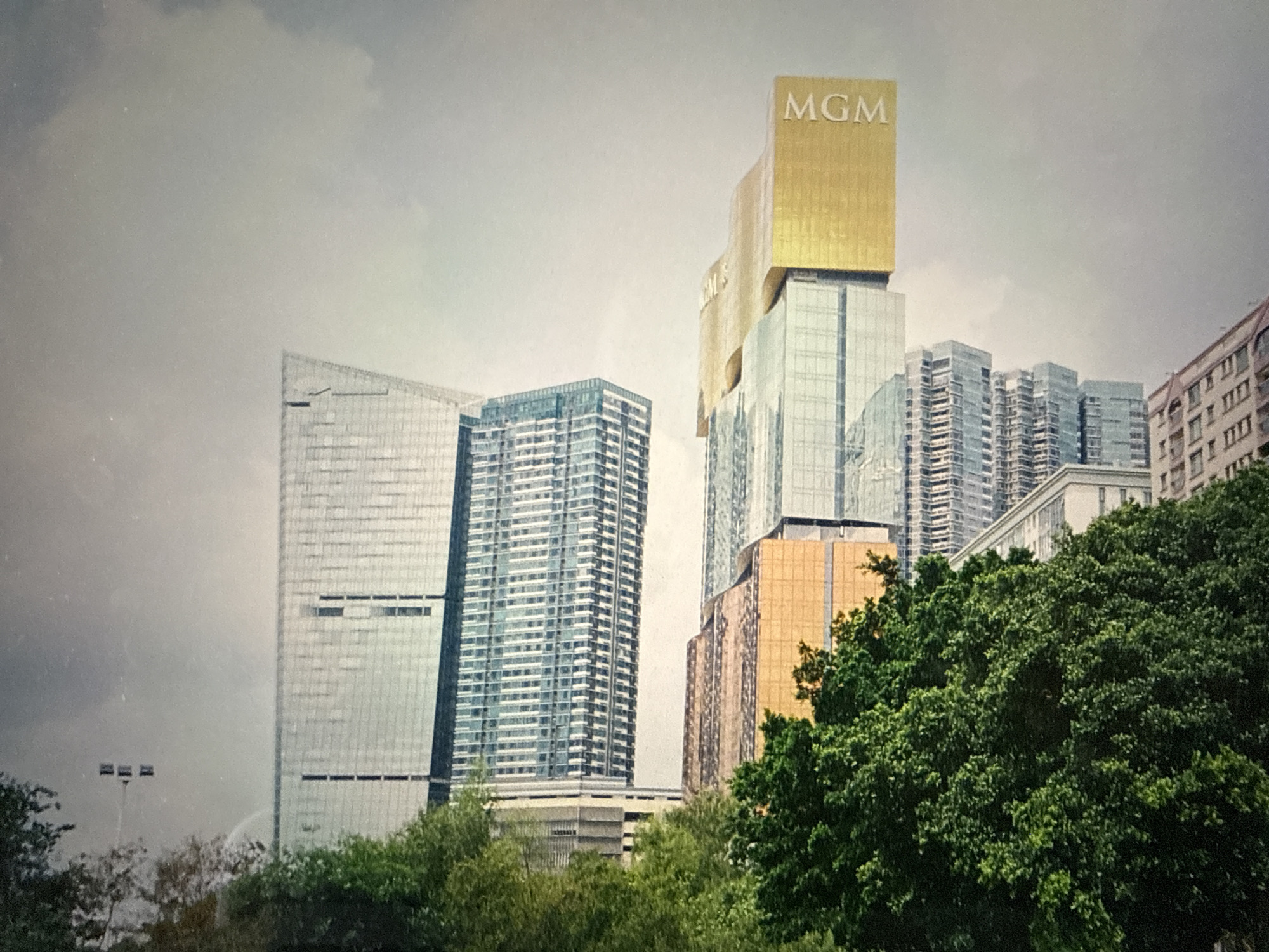 The image shows a group of modern high-rise buildings in an urban setting. The most prominent building is topped with a large gold "MGM" sign. The other buildings are mostly glass and steel, with varying designs and heights. There are green trees and bushes in the foreground, partially obscuring the lower parts of the buildings. The sky is overcast. Based on the architecture, it is highly improbable that the location is Almaty, Kazakhstan. The buildings appear to be casinos and resorts, which are not common in Almaty.