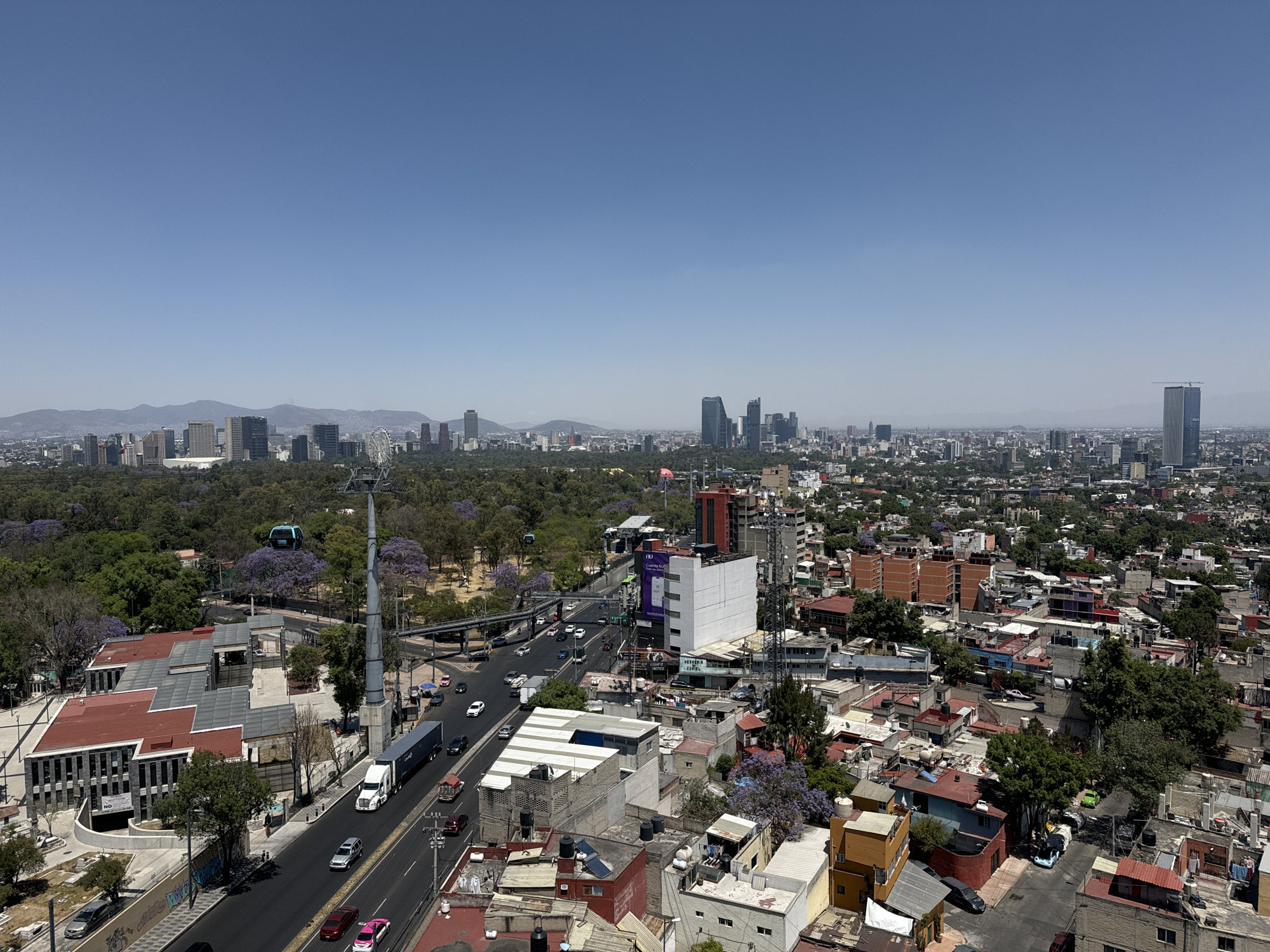 An elevated, wide-angle photograph captures a bustling urban landscape under a clear blue sky, identified as Mexico City, Mexico, specifically from a vantage point in the Iztapalapa borough looking west.
In the immediate foreground on the left, a large, multi-section building with a reddish-brown roof and grey facades, partially under construction or renovation, features a curved access ramp and a banner displaying "NO." Adjacent to this, a multi-lane asphalt road carries traffic including a white semi-truck with a blue trailer, several cars (silver, red, white, pink, black), and an SUV. A prominent, tall grey pylon of the Cablebús (cable car system) stands beside the road, with a blue cable car cabin visible in transit over a large green park area. Numerous Jacaranda trees with purple blossoms are interspersed throughout the park and surrounding areas.
To the right of the main road, the scene transitions into a dense urban fabric of varying residential and commercial structures with diverse roof colors (red, brown, grey, white, orange). A white commercial building prominently displays a digital billboard for "NU," and another structure exhibits "MERCERIA CURIEL" signage. A metal pedestrian bridge or overpass crosses above the main road in the midground.
The background presents an expansive cityscape defined by numerous buildings, ranging from low-rise structures to a distinct cluster of modern high-rise skyscrapers in the mid-right. A tall, slender skyscraper with a construction crane on its apex is visible in the far right distance. A large Ferris wheel-like structure is discernible among the buildings in the left-center background. Distant mountains form a hazy silhouette on the horizon, defining the city's perimeter. The overall scene depicts a vibrant, densely populated metropolitan area under bright daylight conditions, suggesting spring due to the blooming Jacaranda trees. No individual people are distinctly visible or identifiable.