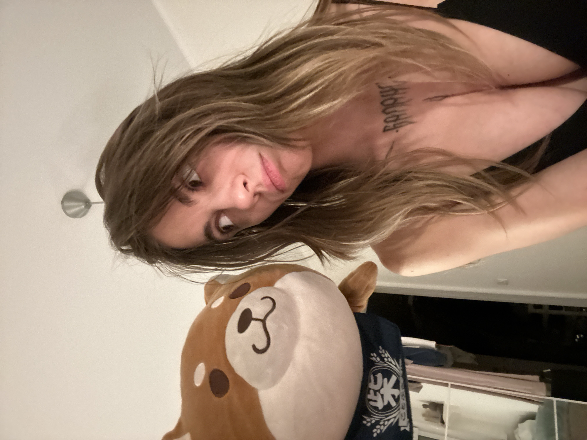 The image shows a young woman indoors, possibly in an apartment. She has long, wavy brown hair and is looking towards the left of the frame with a slightly pensive expression. A plush toy, resembling a Shiba Inu dog wearing a blue bandana with Japanese characters, is positioned to her left and slightly in front of her. The woman is wearing a black top that reveals a tattoo on her chest. The background is out of focus, but appears to be a domestic setting with some clothing items hanging in the background. There is no indication that this image was taken in Moscow, Russia.