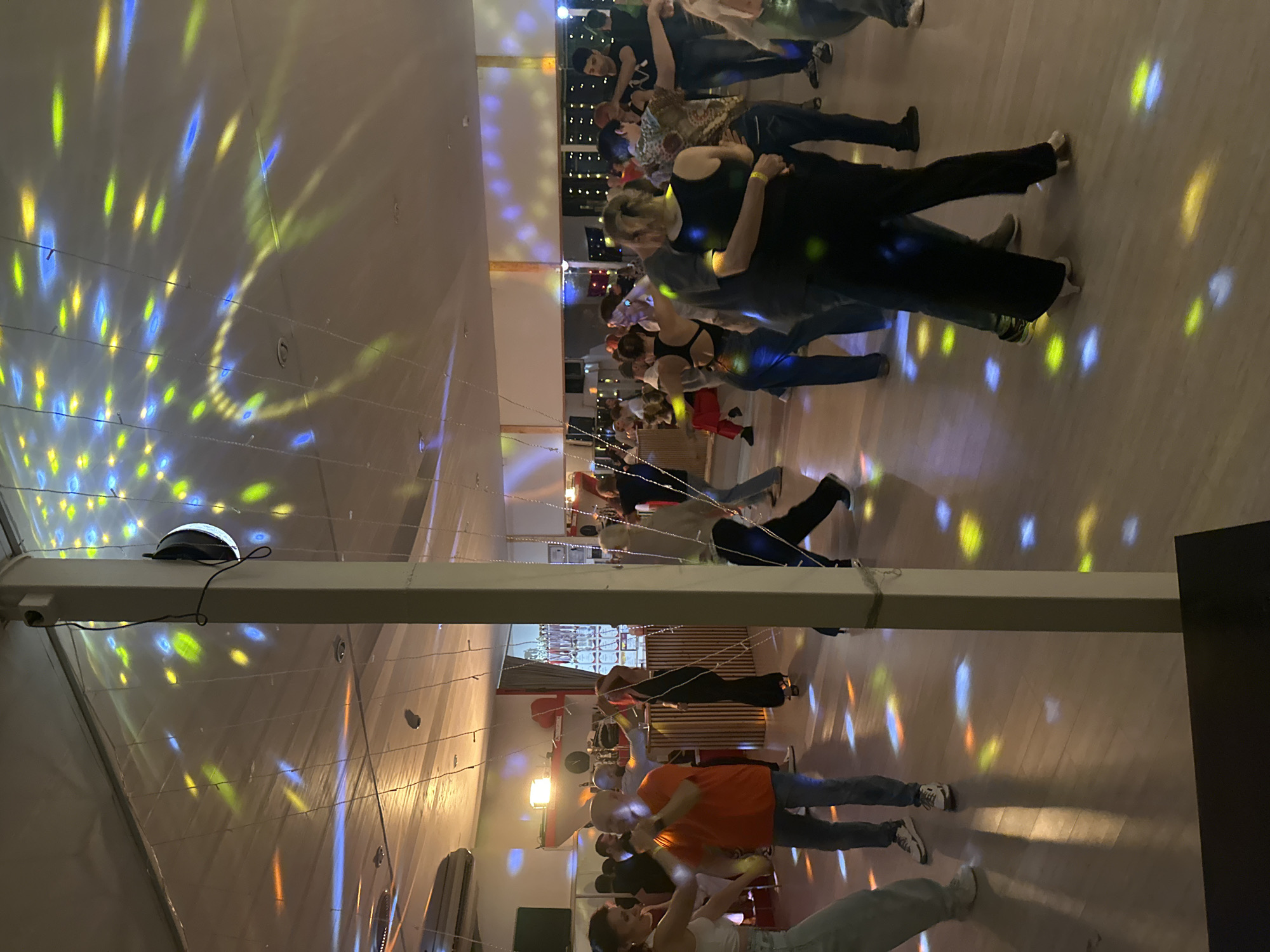 A dimly lit indoor dance floor is occupied by numerous couples, seemingly participating in a dance event. The lighting primarily consists of colorful projections, possibly from a disco ball or similar device, scattered across the walls, floor, and participants. The setting appears to be a social gathering or dance class, with people of various ages and attire engaged in partnered dancing. A supporting beam runs across the top of the image. The event's location in Sochi, Russia, cannot be verified from the image.