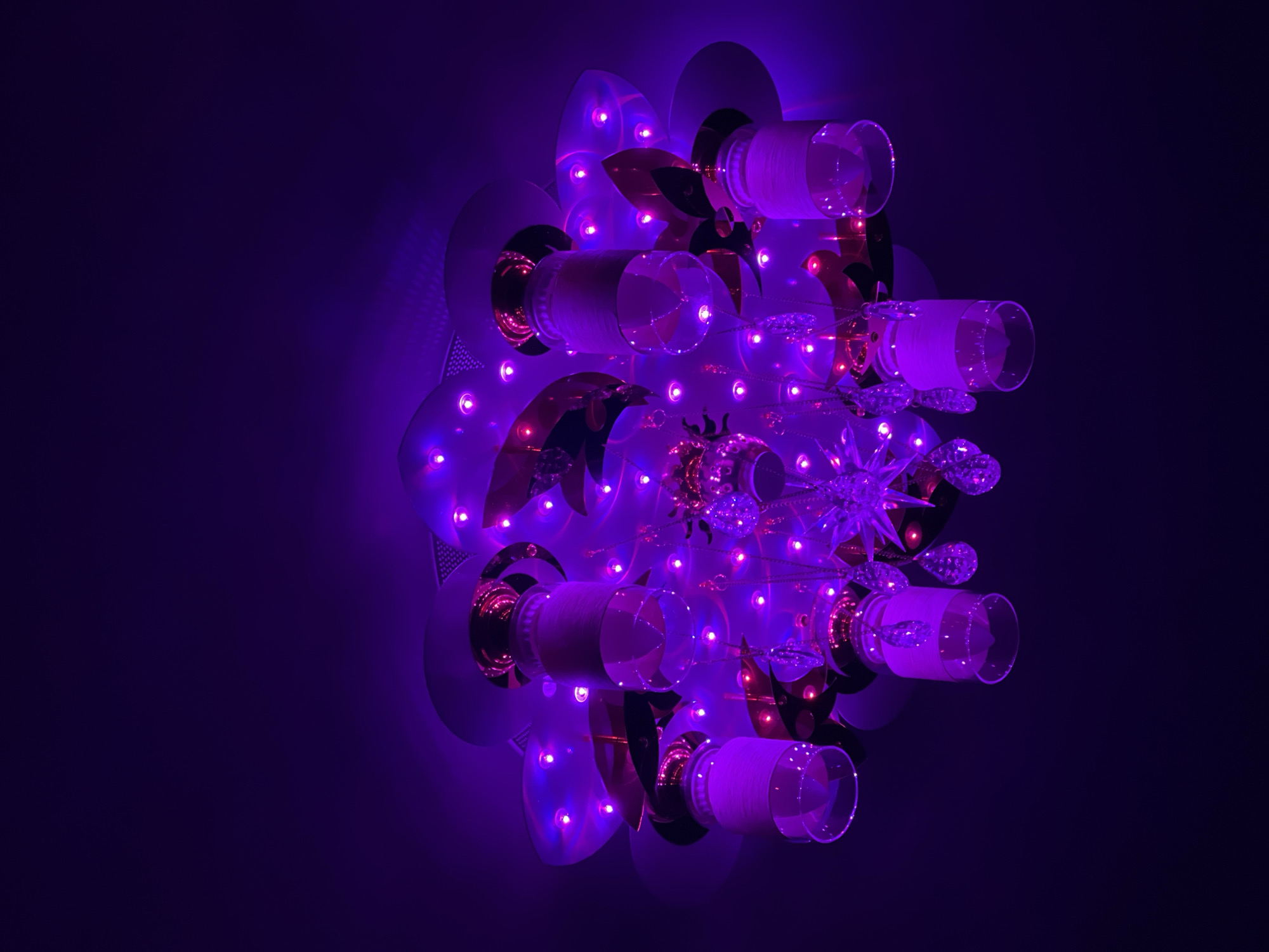 A close-up, low-angle shot captures a decorative lighting fixture against a dark violet background. The fixture has a circular design with clear cylindrical glasses projecting outwards, each with a slightly frosted appearance. Between the glasses are ornamental details, including metallic gold-colored petal-shaped accents and smaller, decorative crystal-like elements. Small purple LED lights are embedded throughout the fixture, providing illumination and highlighting the individual design elements. The overall lighting is soft and creates a surreal, futuristic aesthetic. The location is unknown, Unknown.