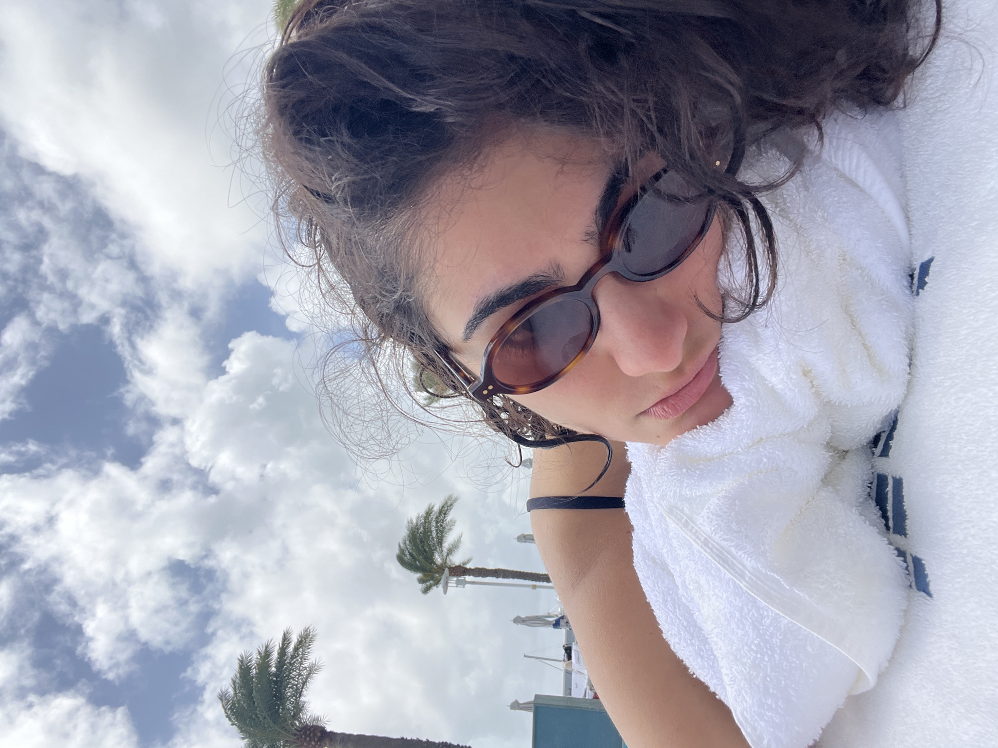 A woman with dark curly hair and sunglasses is lying on a white towel in an outdoor setting, possibly on a beach or near a pool. The sky is visible in the background, with scattered clouds. Palm trees are visible, suggesting a tropical location. Based on these details, the location is likely Miami Beach, United States of America.