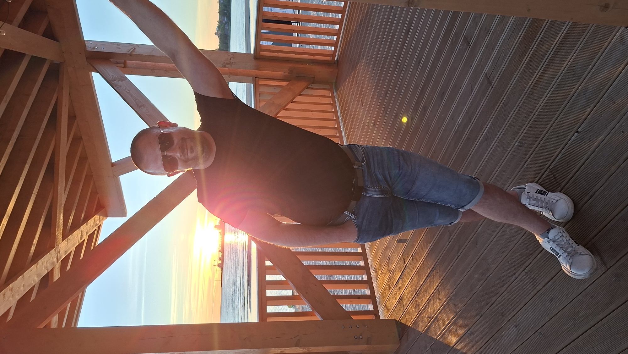 A man wearing a black t-shirt, blue denim shorts, and white sneakers stands inside a wooden structure with a slatted roof. He has sunglasses on and his right arm is extended upwards, resting on a wooden beam. His left hand is in his pocket. The wooden structure appears to be on a deck or boardwalk overlooking a body of water. In the background, the sun is setting over the water, casting a warm glow. A ship or large vessel is visible on the horizon. The overall scene suggests an outdoor setting during sunset, possibly a pier or waterfront area in Tallinn, Estonia, given the characteristic wooden structures and coastal environment.