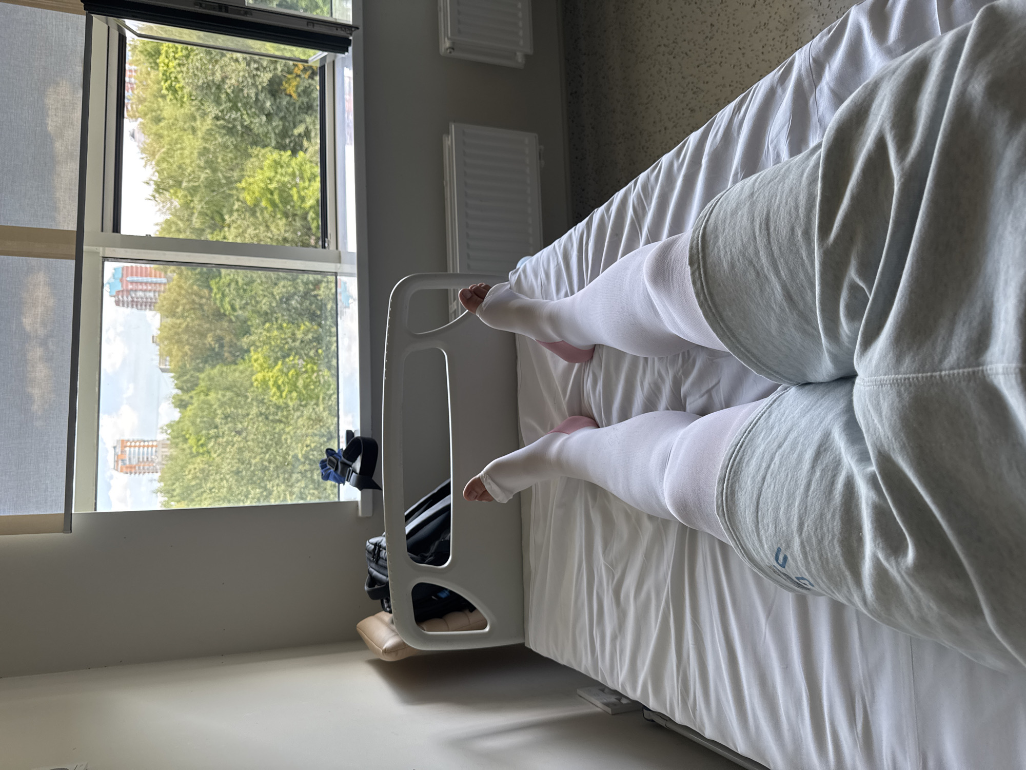 A first-person perspective shot shows a person's legs lying on a hospital bed. The person is wearing compression stockings up to their thighs and grey sweatpants. The bed is covered with white sheets. In the background, a window reveals a cityscape with trees and buildings, suggesting an urban environment. The window also has a roller blind. A hospital bed railing is visible, and to the left, a headrest and some personal belongings, including a backpack and a pair of shoes, are placed on a brown chair. Two radiators are mounted on the wall below the window. The scene is indoors, likely a hospital room, with daylight filtering through the window.