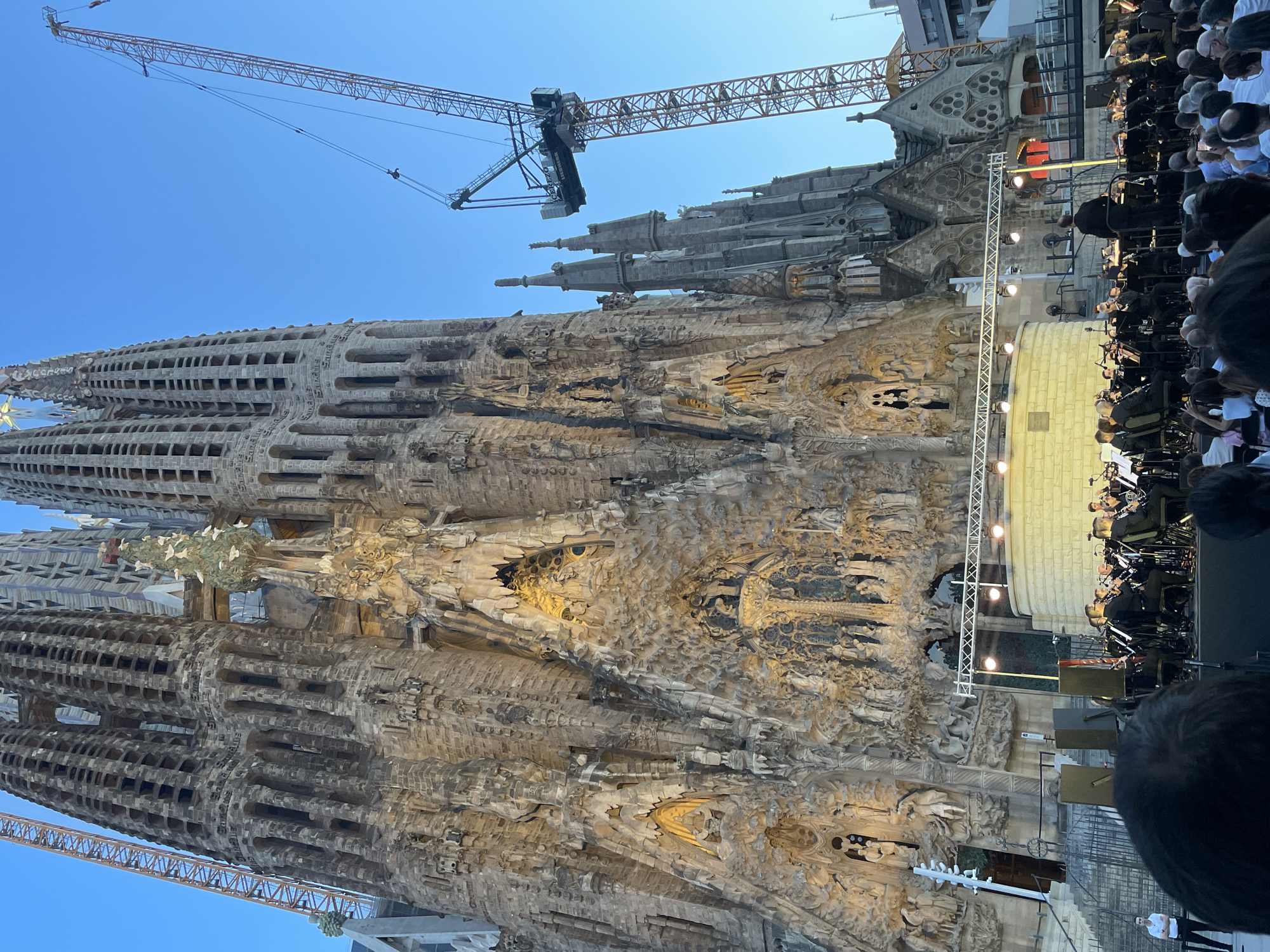 The image depicts an outdoor orchestra performing in front of the Sagrada Familia in Barcelona, Spain. The iconic basilica, a UNESCO World Heritage site, is visible in the background, its intricate facade illuminated by artificial lighting. Two construction cranes are also present, indicating ongoing work on the structure. In the foreground, an orchestra is set up on a stage with lighting rigs, playing for an audience whose heads are partially visible at the bottom of the frame. The sky is a clear blue, suggesting it is daytime or early evening.