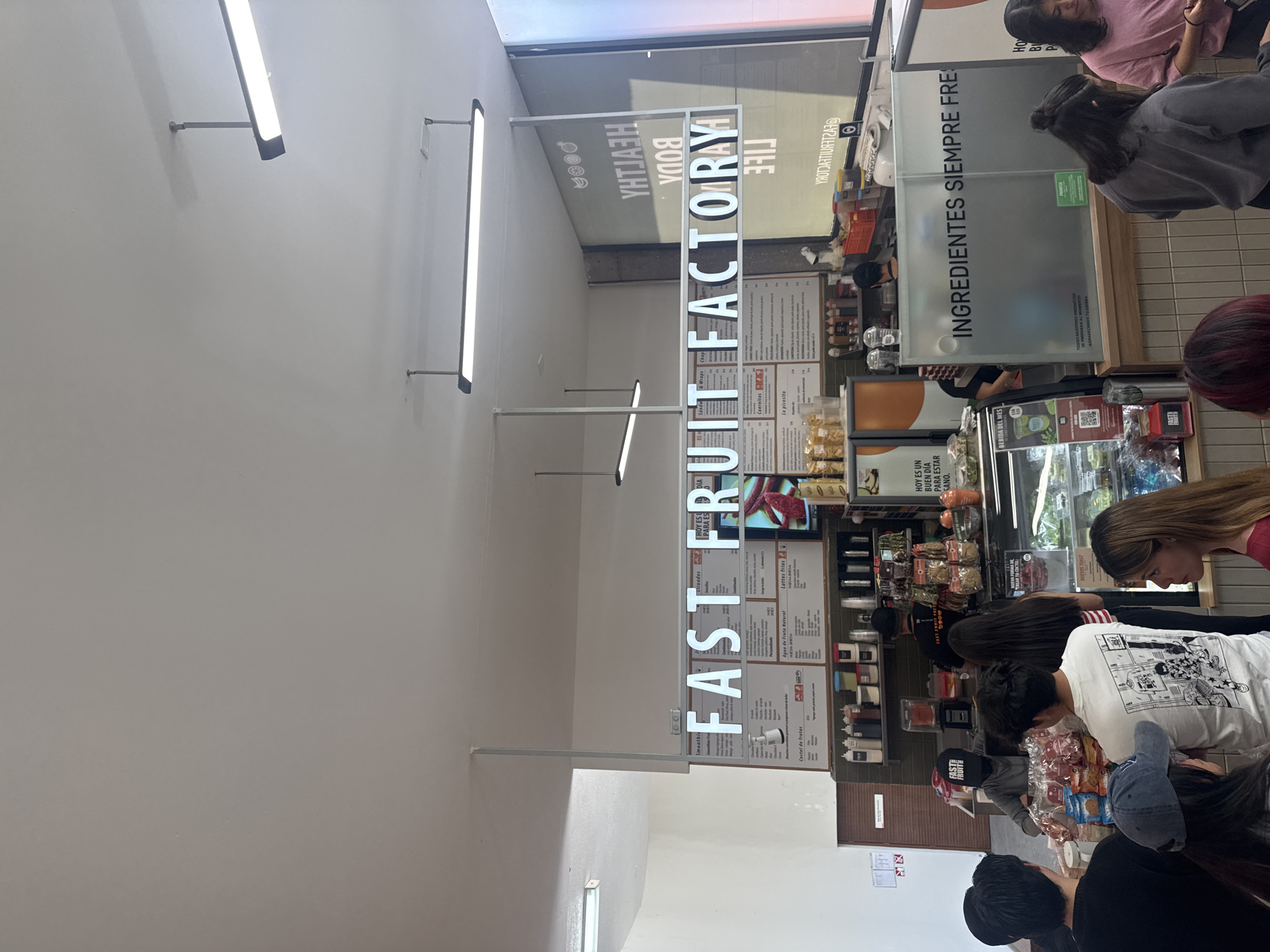 The image shows the interior of Fast Fruit Factory in San Andrés Cholula, Mexico. A brightly lit sign bearing the name of the establishment hangs from the ceiling. Customers are queuing at the counter to place orders, some visible are wearing casual clothing, like t-shirts. The menu board displays various offerings, while food items and condiments are visible behind the counter. The overall atmosphere is that of a busy fast-food restaurant.