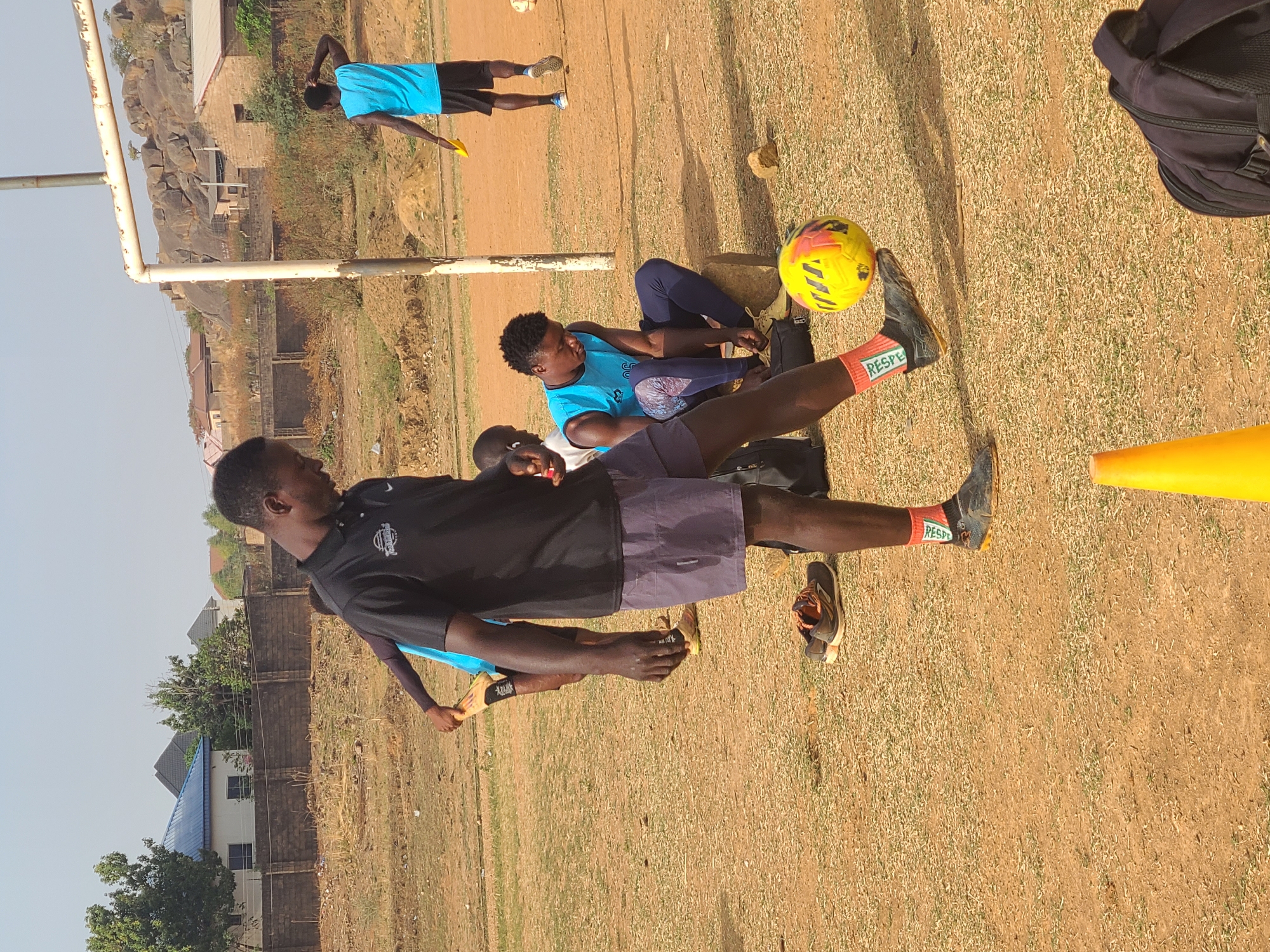 A football training session takes place in Barikin Jirga Summa, Nigeria. There are several young men dressed in athletic wear on a dry, grassy field. One man is standing and appears to be about to kick a yellow football. An orange cone sits to the right of him. A man is sitting behind him, and another man is standing in the background. In the far background, there is a building with a blue roof. A rocky hillside is visible in the background on the left. A football goal is also visible in the background.