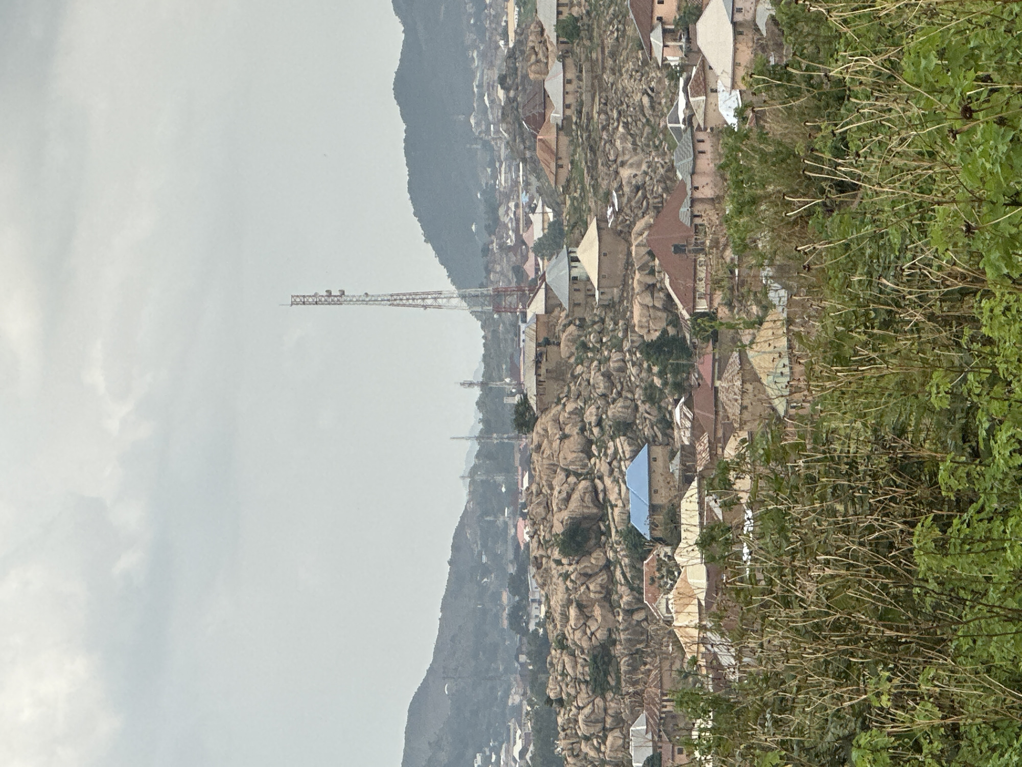 This landscape image depicts a settlement nestled within a rugged, rocky terrain under an overcast sky, consistent with Jos, Nigeria.
In the foreground on the right, dense green foliage and drier, brush-like vegetation dominate the frame. Beyond this, the main scene unfolds, showcasing numerous large, weathered rock formations that define the topography. A town is built around and upon these rocks, with many buildings, primarily houses, visible. These structures feature a mix of roof types, including reddish-brown tiled roofs, lighter flat roofs, and one prominent blue-colored roof.
A very tall telecommunications tower, constructed with a red and white lattice framework, stands prominently in the middle-left section of the image, stretching into the cloudy sky. Several smaller, similar towers are also discernible on the distant hills. These hills form the backdrop, appearing hazy and muted under the pale, cloudy sky. The weather appears overcast, suggesting diffused light rather than direct sunlight. No people or specific activities are visible within the frame, offering a static view of the urban landscape integrated with its natural, rocky environment. There is no visible text.