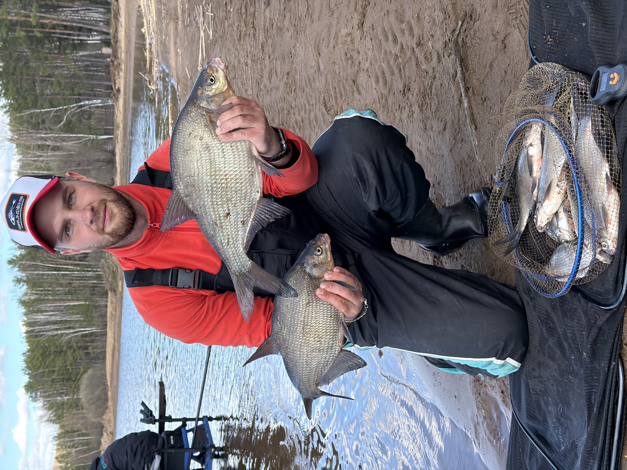 A man is sitting on the bank of a body of water, possibly the Kama River, in what appears to be the city of Chaikovsky, Russia. He is holding two fish in his hands, presumably caught from the river. The man is wearing a red jacket, black pants, and black boots. A fishing net filled with several similar-looking fish is visible to the right of the man. Fishing equipment is visible to his left. The background shows a shoreline with sandy banks and trees.