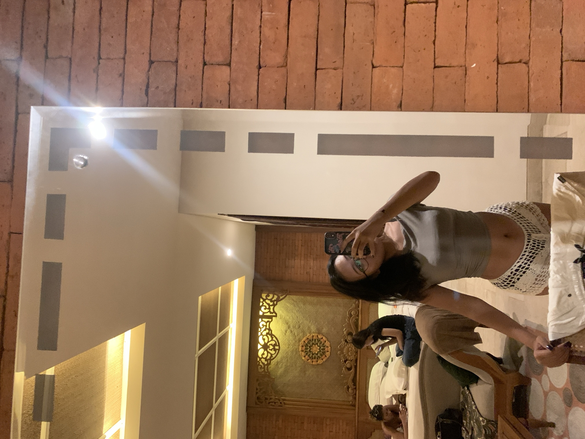 An indoor mirror selfie captures a woman in a grey crop top and white crocheted shorts, holding a smartphone with a floral case to take a picture. She has long dark hair and wears glasses. The image is rotated 90 degrees clockwise, making the brick wall appear as a ceiling and the floor as a side wall.
The large rectangular mirror features matte grey horizontal and vertical stripes as part of its design. Reflected in the mirror, behind the main subject, are two other individuals. One person is sitting on a bed or sofa, wearing a dark top and light-colored pants, engrossed in their phone. Another person is partially visible, appearing to be lying down on the same furniture.
The room's decor suggests a traditional and rustic aesthetic, common in places like Ubud District, Indonesia, which is the provided location context. A prominent feature is an ornate wooden wall panel with intricate carvings, including a central circular, mandala-like motif, set against a woven material. A textured brick wall is visible above the mirror, contributing to the warm, earthy palette of the space.
The setting appears to be a hotel room or guesthouse in the evening or night, illuminated by warm artificial light from various fixtures, including overhead lights and accent lighting along wall panels. The atmosphere is relaxed, with the people present engaged in personal activities. No discernible text is visible on any objects in the image.