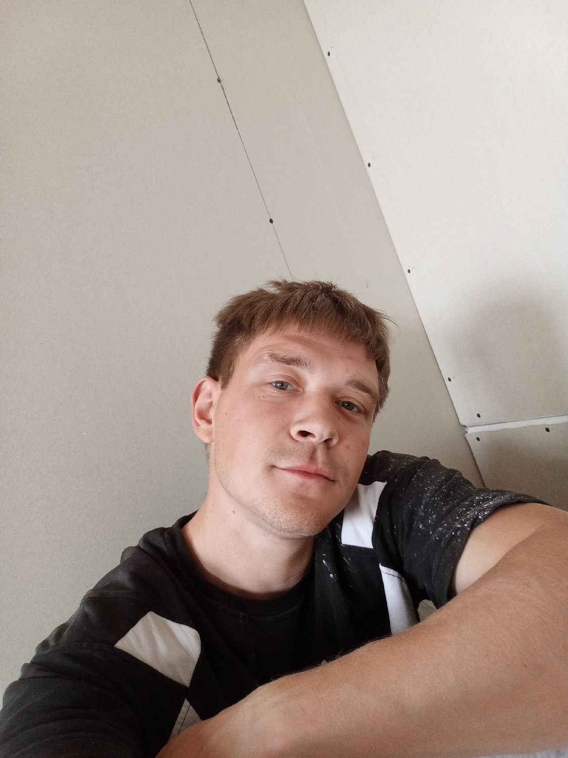 The image displays a selfie of a young adult man indoors in Chaikovsky, Russia. He has short, light brown hair, light-colored eyes, and a neutral, slightly relaxed expression as he looks directly into the camera. He is wearing a black t-shirt with white geometric patterns on the shoulders and upper sleeves. His right arm, with a bare forearm, is visible in the lower right foreground, resting casually.
The background shows a room that appears to be under construction or renovation. The walls are covered with light-colored drywall or plasterboard, featuring visible screws or fasteners embedded in the panels. A thin, dark string or line stretches vertically across the upper left side of the background, likely a plumb line used for alignment in construction. The wall on the right side of the frame is noticeably angled or sloped, suggesting an attic space or a room with a pitched ceiling. The lighting is consistent with an indoor setting, and no specific time of day is discernible. No visible text is present in the media.