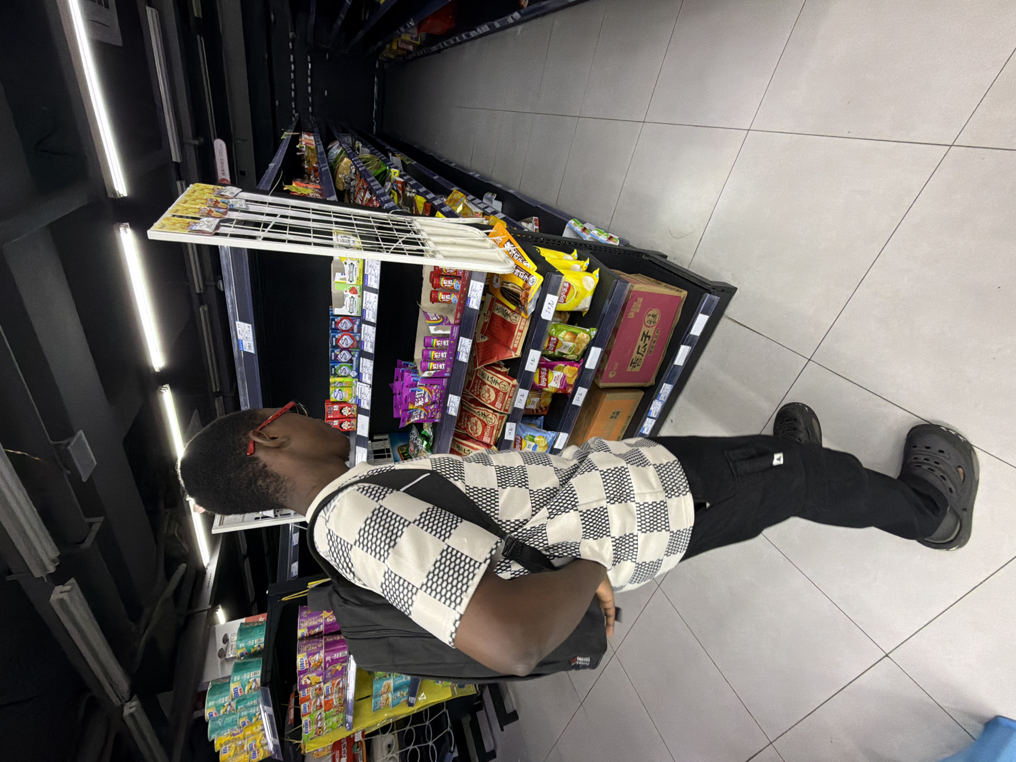 A Black male wearing a black backpack, a black and white checkered shirt, black cargo pants, and black clogs is standing in a brightly lit grocery store aisle, looking at a display of snacks. The store appears to be located in Jiangjin, China, based on the product packaging visible in the image, which includes Chinese characters. The shelves are stocked with various food items, including chips, candies, and boxed snacks. The aisle is clean with white tiled flooring. The man is positioned in the center of the frame, with the shelves extending into the background on both sides. The overall impression is that of a typical shopping scene in a modern supermarket.
