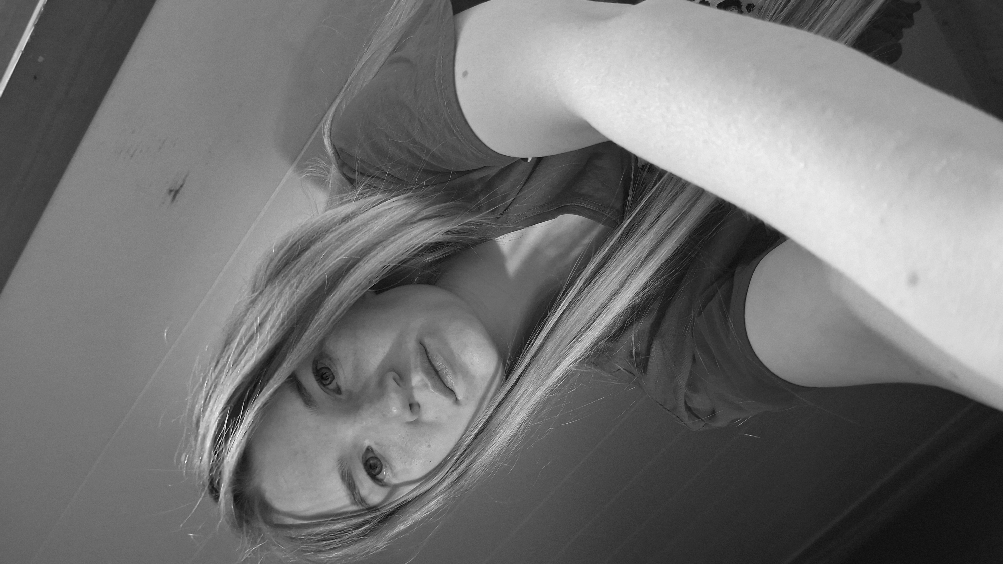 The black and white photograph shows a close-up, low-angle view of a young woman from the shoulders up. She has long, blonde hair that falls forward, partially obscuring her face. Her eyes are open and looking towards the camera. She is wearing a dark t-shirt. Her right arm is extended forward, taking up a significant portion of the foreground. The background appears to be an indoor wall with horizontal lines and some smudges or marks. There is no indication of the city of Большой Букор, Russia within the image.