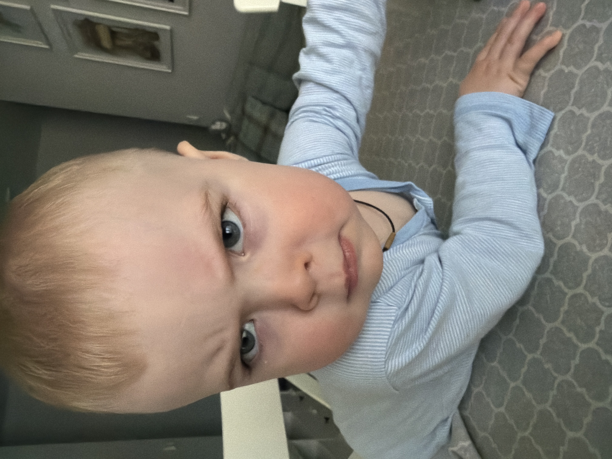 The image presents a close-up, top-down view of a baby or toddler, likely taken indoors in Votkinsk, Russia. The child, with fair skin, light blonde hair, and blue-grey eyes, is positioned diagonally in the frame while lying down. They wear a light blue and white striped long-sleeved shirt. A thin, dark cord necklace with a small, light-colored bead or pendant is visible around their neck. The child has rosy cheeks and a somewhat serious or curious expression, gazing directly at the camera with wide eyes. Their left arm is extended towards the right side of the frame. The immediate background beneath the child is a grey surface with a repeating, geometric pattern, possibly a bedspread, rug, or playmat. Further in the background, out of focus, framed pictures can be seen on a wall, indicating a room setting such as a bedroom or nursery. The lighting suggests it is daytime. There is no visible text in the image.