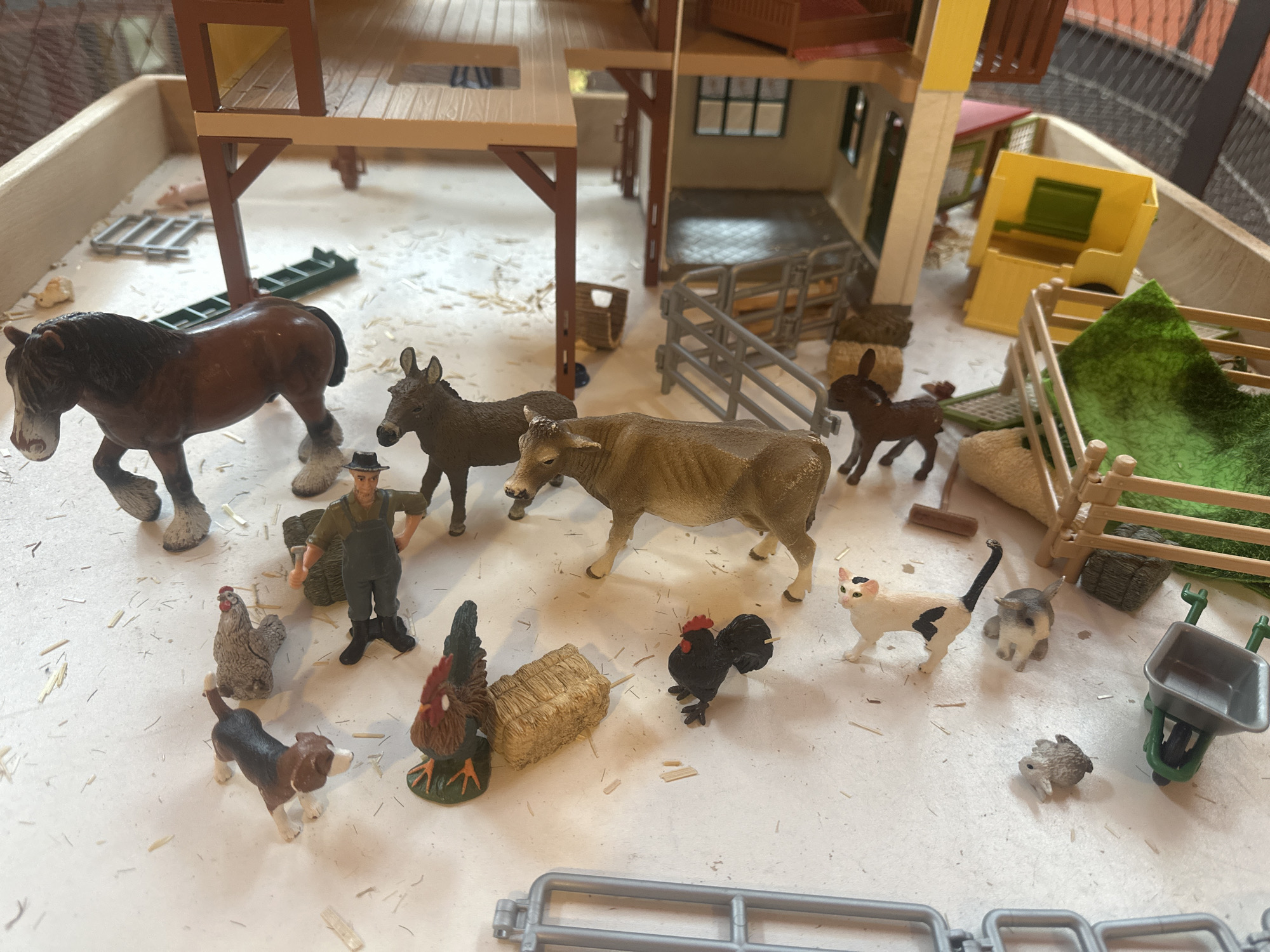 In a toy farm set, a variety of plastic animal figures and accessories are arranged on a light-colored surface scattered with small straw-like pieces, simulating a farm environment. In the foreground, a brown and white dog, a rooster with a brown and red body on a green base, a small bale of hay, and a black rooster are visible. To the left, a light brown and gray chicken is positioned near the dog. A farmer figure in green overalls and a hat stands next to a bale of hay. In the midground, a brown horse stands to the left, followed by a donkey, a light brown cow, a dark brown calf, and a black and white cat. A gray rabbit is situated to the right of the cat, with another smaller gray animal behind it. A small gray and white cat is visible behind the calf. A red and green toy wheelbarrow is located on the far right. In the background, a multi-story toy barn structure with open floors and railings is prominently featured, with a small pink pig figure visible on a lower level. Various fencing elements and hay bales are also part of the scene, along with a yellow and green toy structure resembling a building or equipment. The overall scene depicts a miniature farm setting with numerous toy farm animals and accessories.