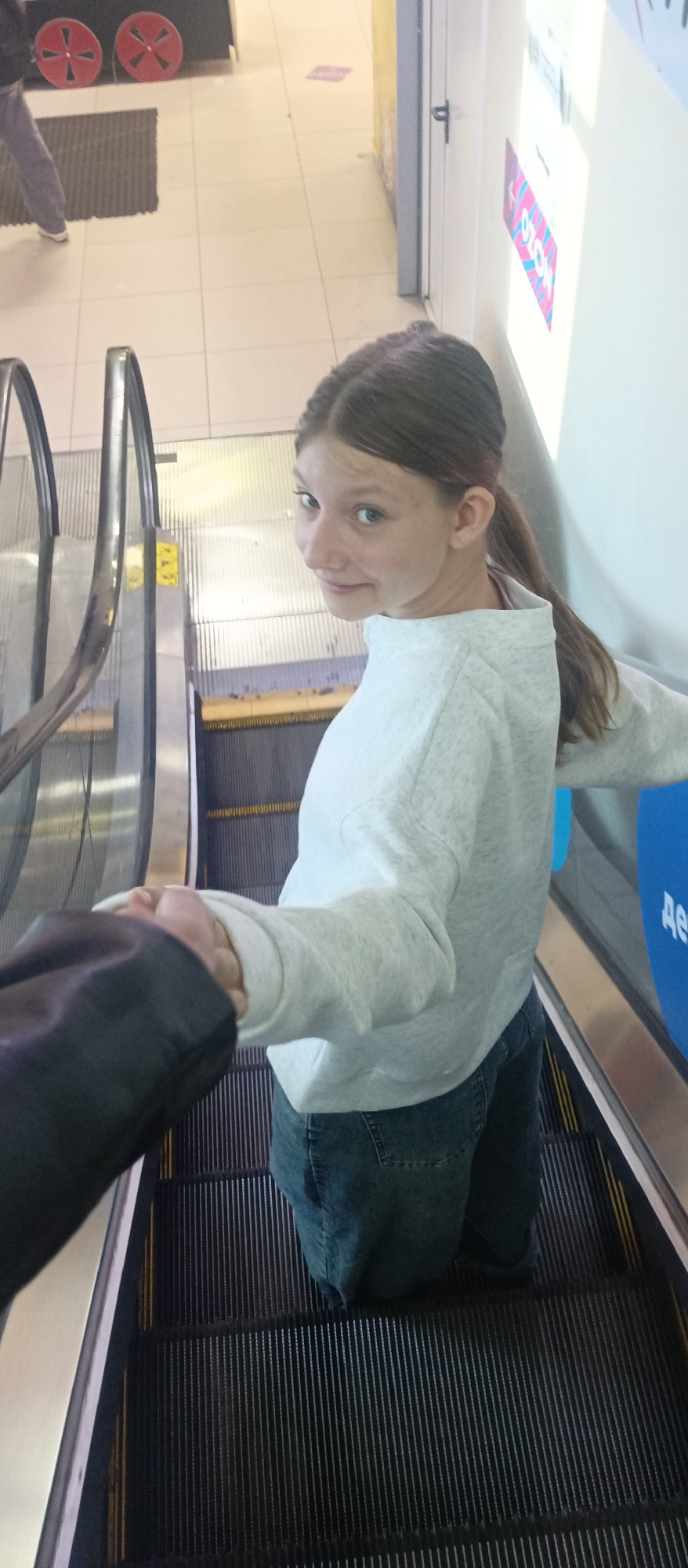 A young person is on an escalator in an unknown location, possibly a shopping mall. They are holding hands with someone whose arm is visible on the left. The person is wearing a light grey sweatshirt and dark wash jeans, and their hair is pulled back in a ponytail. They are looking over their shoulder at the camera. The escalator is moving upwards. In the background are a white wall with a sign, and flooring.