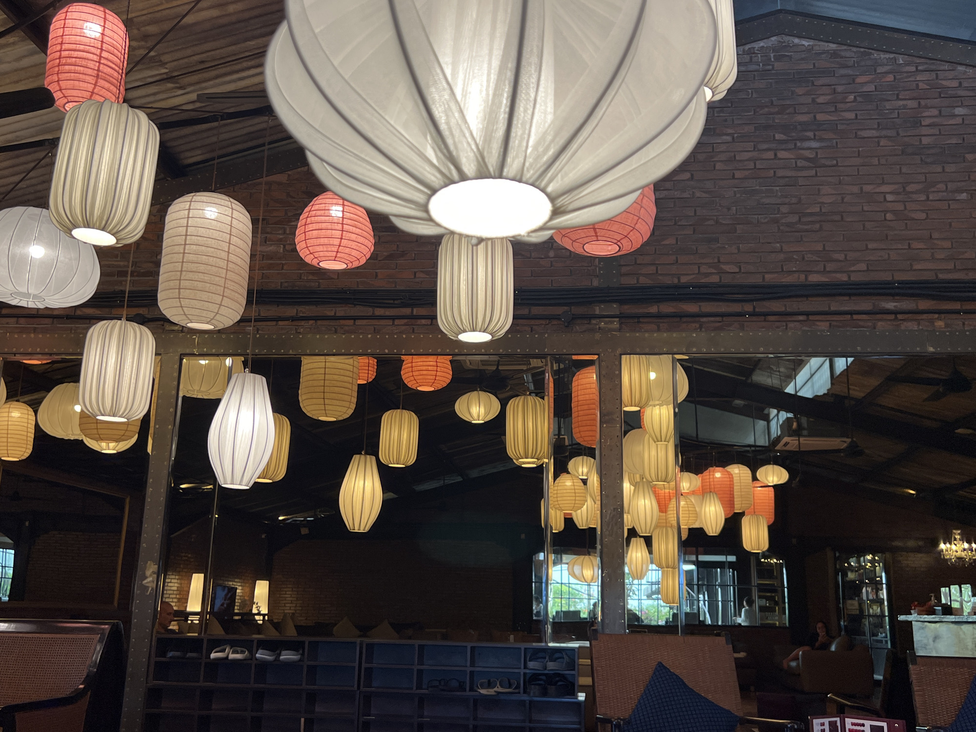 The image depicts an interior scene in North Kuta, Indonesia, featuring a cafe or restaurant setting characterized by numerous hanging lanterns of various shapes and colors, including red, white, and beige. The lanterns are suspended from a dark ceiling with visible wooden beams and exposed brick walls. The foreground shows parts of seating arrangements, including rattan chairs and cushioned seating. In the mid-ground, a large mirror reflects the lanterns and the interior space, revealing more seating areas and a shoe rack with footwear. The background includes a bar area with a chandelier and a window with a view of the outdoors. There are a few individuals visible in the background, appearing to be patrons or staff. The overall atmosphere suggests a dimly lit, cozy environment.