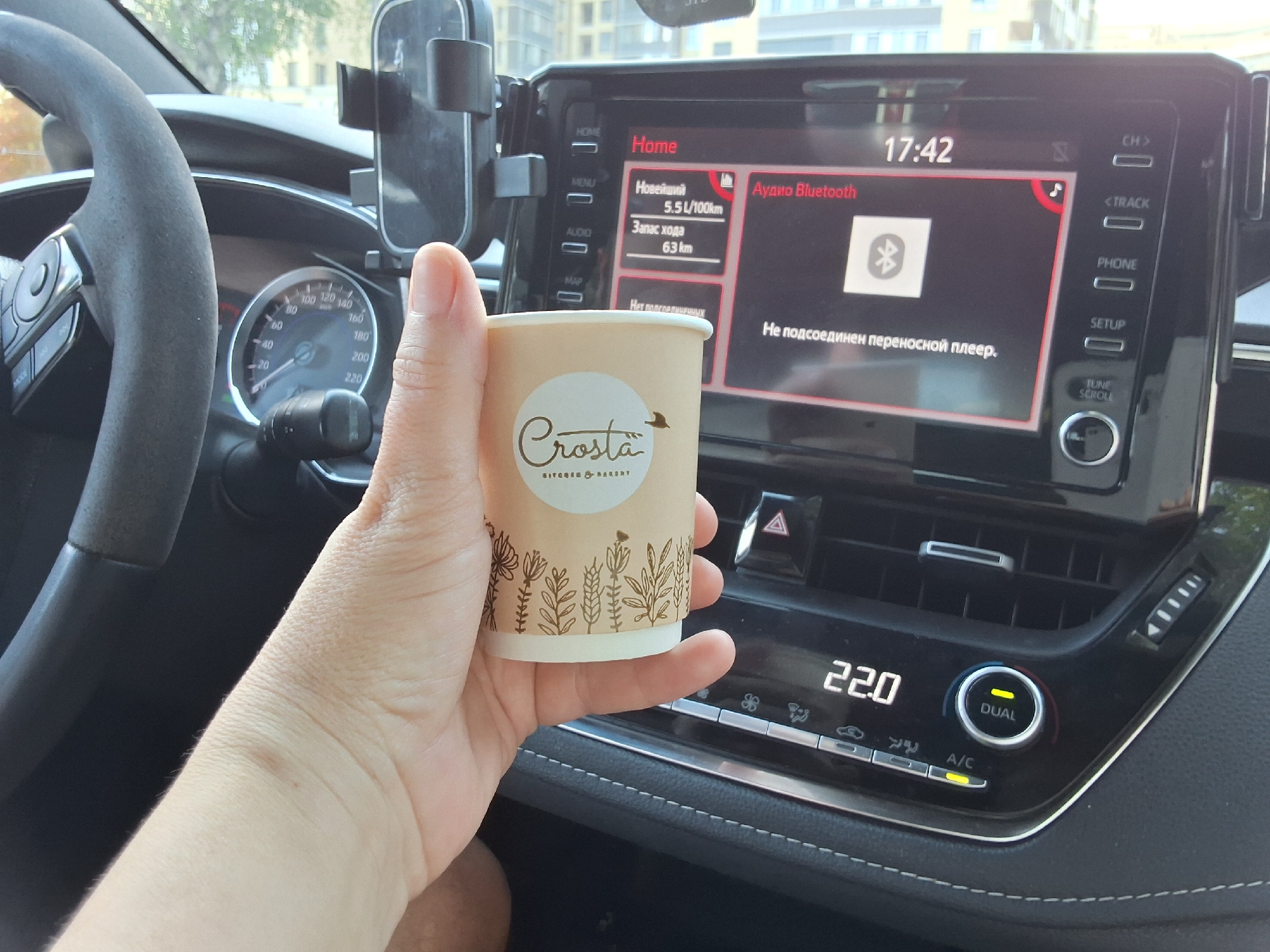 In an interior shot taken from the passenger seat of a car, a person's hand holds a disposable coffee cup with a floral design and the logo "Crosta KITCHEN & BAKERY". The car's dashboard is visible, featuring a digital display showing the time as "17:42" and a car infotainment screen. The infotainment screen displays "Home", "Audio Bluetooth", a Bluetooth icon, and the text "Не подключен переносной плеер." (No portable player connected). To the left of the infotainment screen, a vehicle's speedometer is partially visible. The car's interior includes a steering wheel on the left and climate control buttons on the right, with a temperature display reading "22.0". The scene is illuminated by natural daylight, suggesting it is daytime.