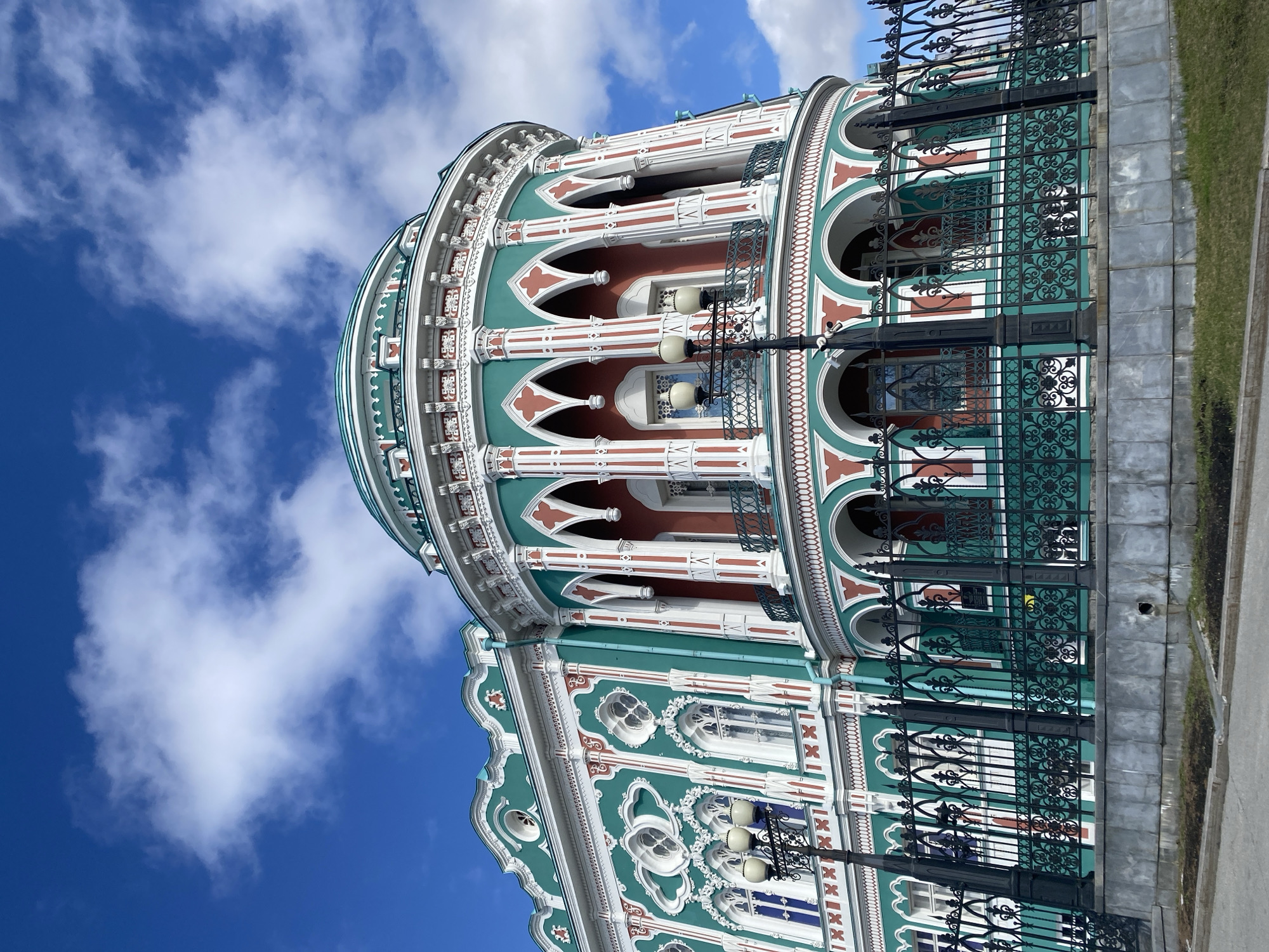 The image shows the exterior of the Sevastianov House in Yekaterinburg, Russia. The building exhibits a distinctive architectural style, featuring multiple tiers adorned with white columns, arched windows, and decorative green and red accents. An ornate black iron fence runs along the base of the building. The sky is blue with scattered white clouds. The angle of the photo is from a low vantage point, emphasizing the building's height.