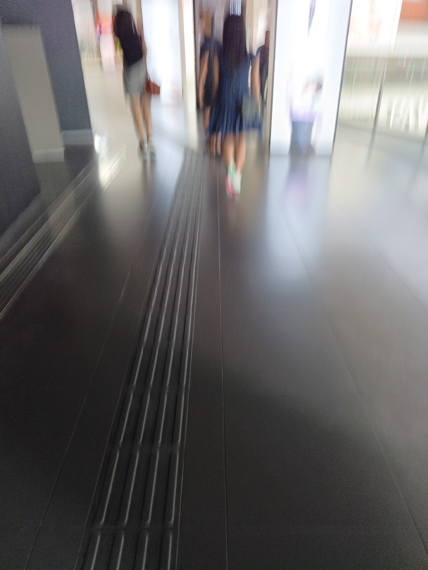 The image shows a blurred scene of people walking in an indoor environment, possibly a shopping mall or a transit station. The floor is dark and reflective, with a tactile paving strip running across it. In the foreground, the textured tactile paving is visible, consisting of raised, parallel bars designed to assist visually impaired individuals. Further in the background, two individuals are walking away from the camera. One person on the left wears a dark top and a light-colored skirt or shorts, carrying a bag over their shoulder. The other person on the right wears a dark dress and colorful shoes. The environment appears to be brightly lit, with soft lighting reflecting off the floor. The blurriness suggests motion, possibly from the camera or the people moving. Based on the distinctive tactile paving pattern, which is commonly used in Malaysian public spaces, and the general architectural style, this scene is likely located in Kuala Lumpur, Malaysia.