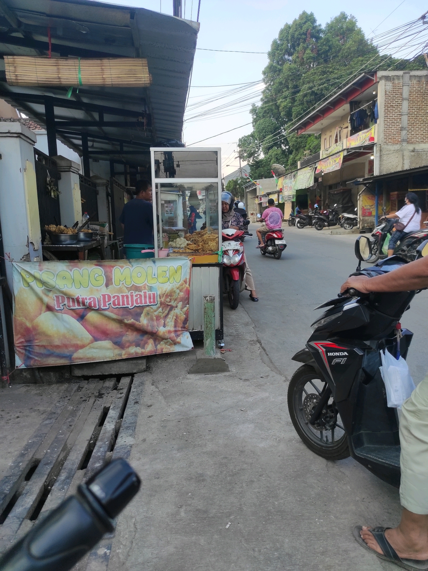 In the city of Padalarang, Indonesia, on what appears to be a street with moderate pedestrian and vehicle traffic, a food stall selling "Pisang Molen" (a type of fried banana fritter) is prominently displayed. A large banner with the text "PISANG MOLEN Putra Panjalu" is hung across the front of the stall. Behind the counter, a man in a dark blue t-shirt is visible, seemingly attending to the food. The stall has a clear glass display case showcasing the fried goods, and several metal containers are present, likely holding more food items.
Several motorcycles are present in the scene. One black Honda motorcycle is in the foreground, positioned at an angle to the viewer, with its front wheel and handlebars visible. A person's hand is resting on the handlebars. Another red motorcycle with a rider wearing a pink shirt is further down the street. A white motorcycle with a rider wearing a white shirt and patterned pants is also visible in the background, as are other parked motorcycles.
The environment suggests an urban setting, with buildings lining the street. Some buildings have balconies with laundry hanging out to dry, and various signs and banners are attached to their facades. Electrical wires are strung across the scene, indicating the presence of infrastructure. The ground is paved with concrete, and a wooden grate or drain cover is visible in front of the food stall. The overall lighting suggests it is daytime, possibly late afternoon or early evening, with the sun casting some shadows.