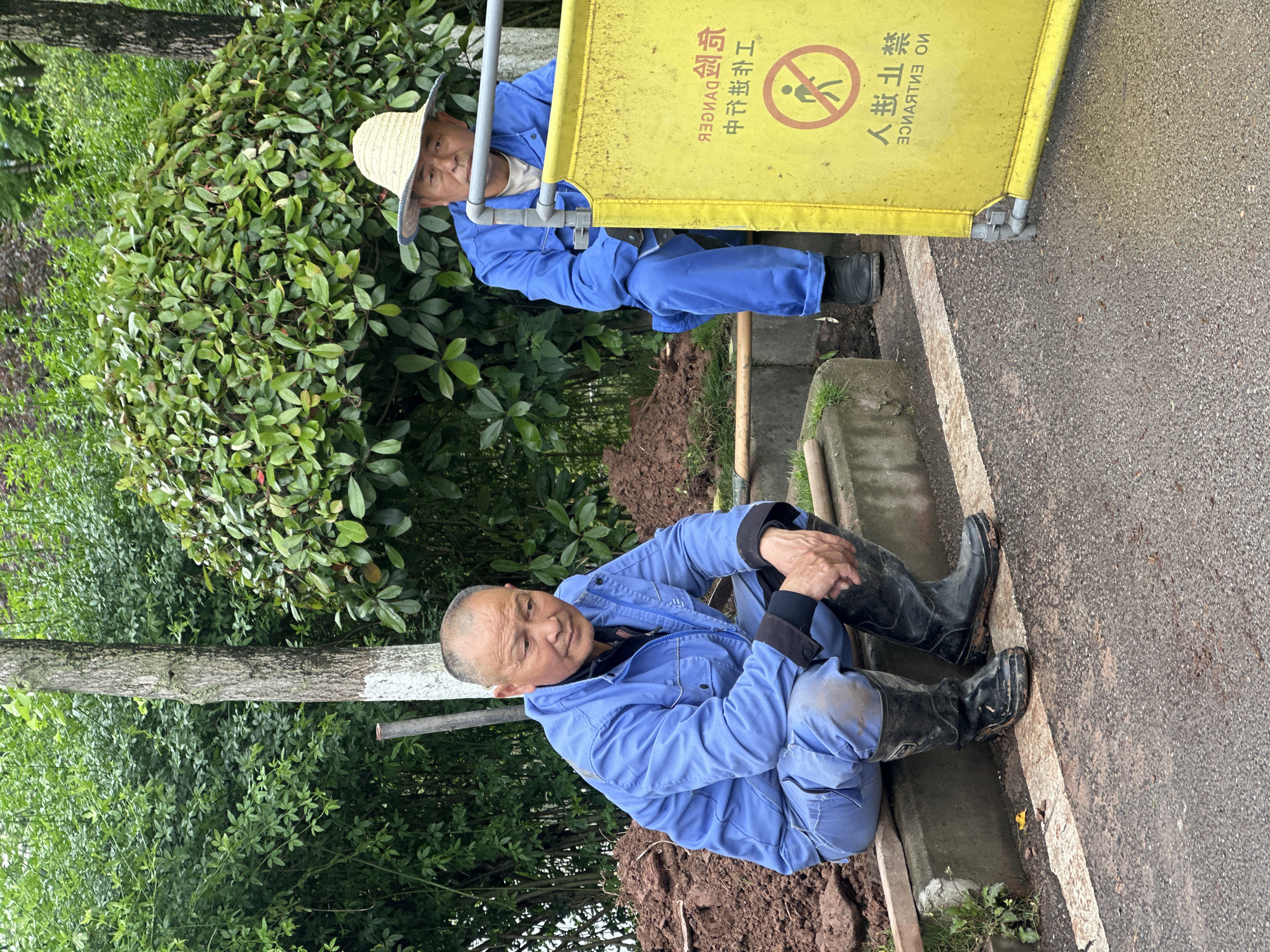Two middle-aged Asian men are working in Jiangjin, China. One man is seated on the ground near a freshly dug area, wearing a blue uniform and black boots. He is looking towards the camera. Another man, also in a blue uniform and a straw hat, is seen further back, possibly working with a yellow barrier sign indicating "No Entrance" in Chinese characters. The environment includes green foliage, a tree, and a paved area.