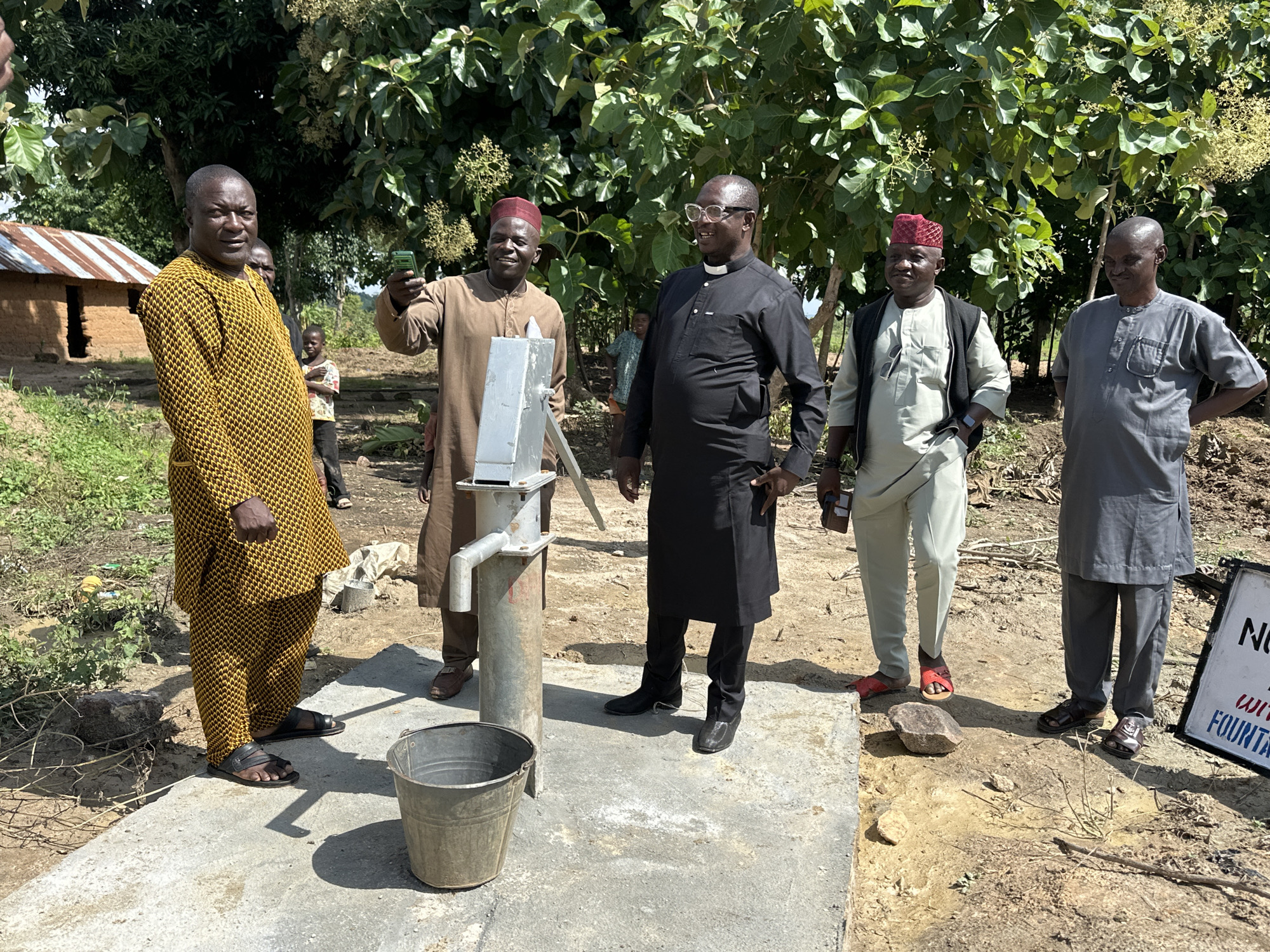 In the city of Afogo, Nigeria, a group of five men stand near a water pump. The man on the left wears a yellow and black patterned tunic and pants. The man next to him, wearing a tan outfit and a red cap, holds up a cell phone, possibly taking a photo or video. To his right, a man in a black cassock with a white clerical collar smiles, and next to him, a man in a light-colored tunic and vest stands with his hands on his hips. The man on the far right wears a grey tunic and pants. A metal bucket sits beside the water pump, which is situated on a concrete platform. In the background, a small mud-brick house with a corrugated metal roof is visible, along with trees and dry, dusty ground.