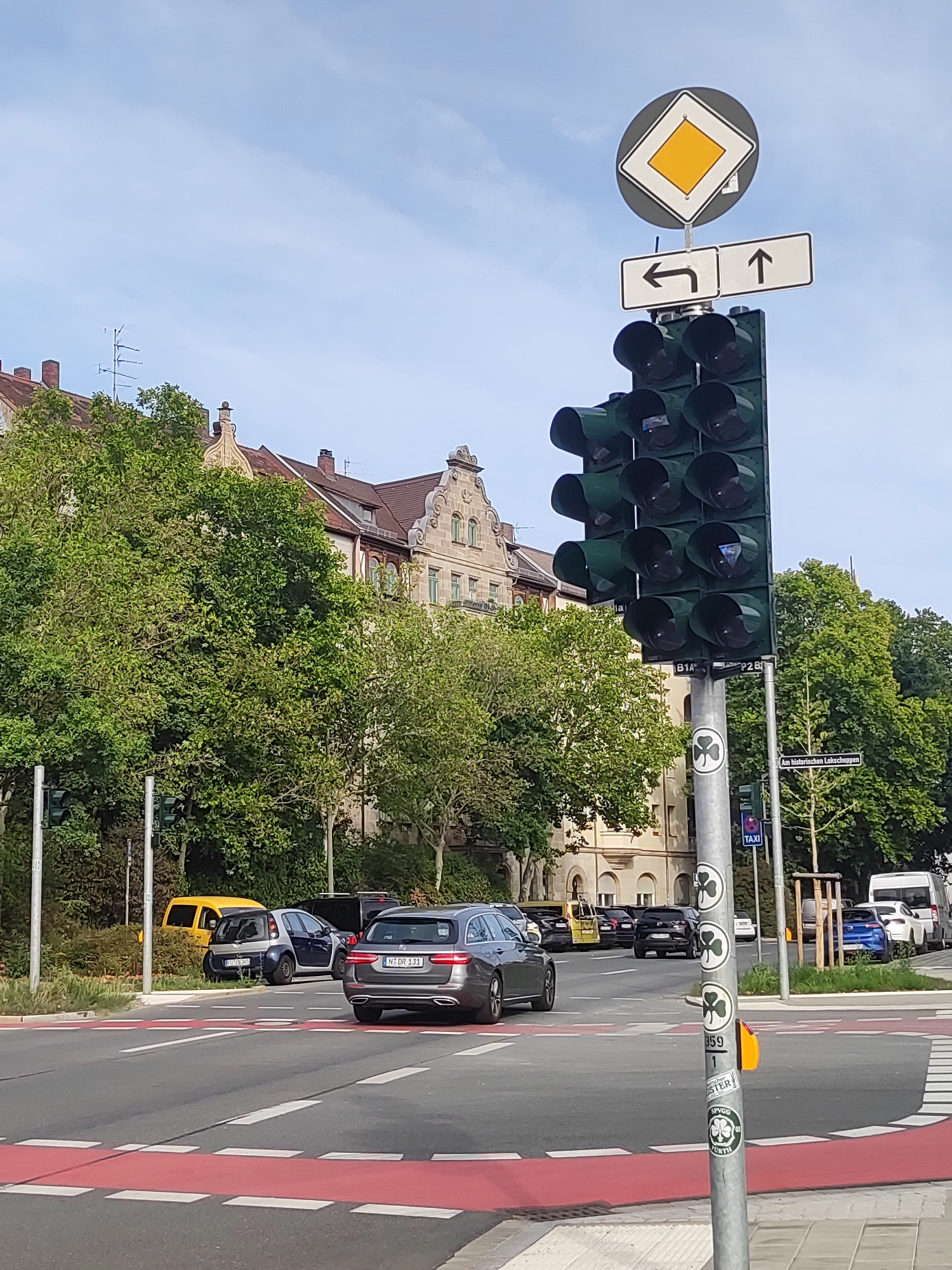 In Fürth, Germany, on a clear day with a blue sky, a traffic light stands tall at an intersection. The traffic light displays multiple dark green lenses, indicating it is not currently active with any illuminated signals. Above the traffic light, a diamond-shaped sign with a yellow center is visible, accompanied by two rectangular signs. The left sign depicts a left-pointing arrow, and the right sign shows an upward arrow.
On the street, several cars are present. A gray Audi is in the foreground, facing away from the camera. To its left, a small dark blue and silver car is parked, and behind it, a yellow van is also parked. Further down the street, other vehicles are seen, including a dark SUV, a white van, and several other cars.
The background features buildings with traditional European architecture, characterized by red-tiled roofs and ornate facades. Lush green trees line the street, contributing to the urban landscape. A street sign indicates "Am historischn Lekschnuggen," and a "TAXI" sign is also visible on a pole. The intersection includes a crosswalk with red and white markings. A pedestrian crossing button is mounted on the traffic light pole. Several stickers with shamrock imagery are affixed to the pole.
