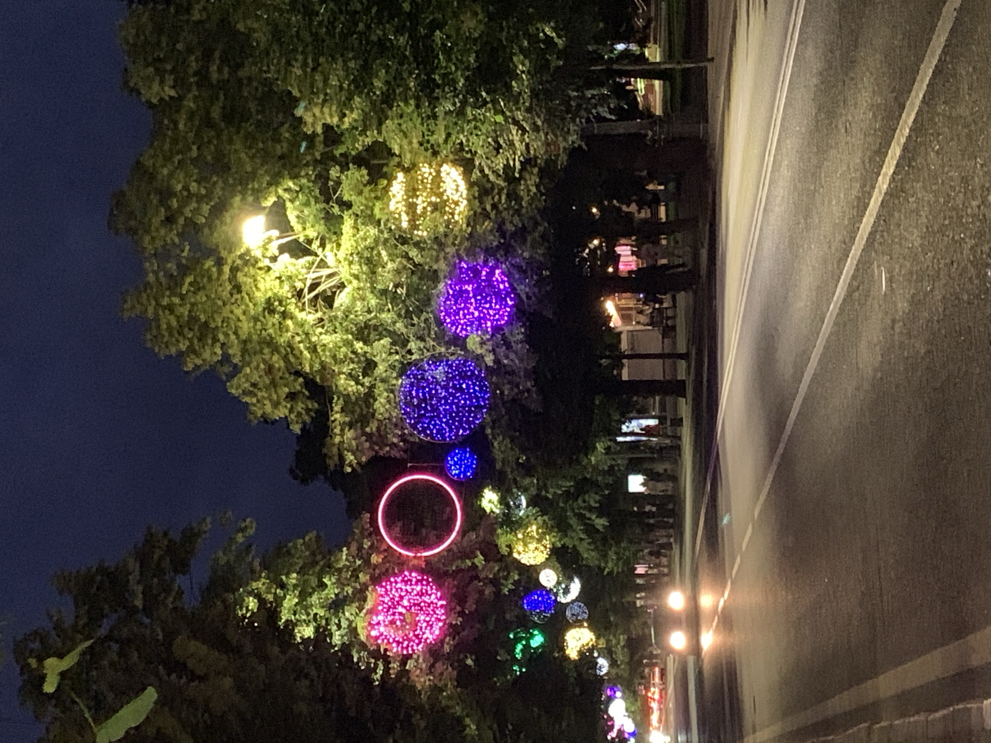 An eye-level, medium shot captures a street at night decorated with colorful spherical light installations hanging from trees. The road, a two-lane asphalt surface with visible yellow and white markings, is illuminated by the headlights of oncoming vehicles and streetlights. Several cars are driving on the road, their headlights creating bright streaks of light. The trees lining the street are adorned with various colored lights, including pink, purple, blue, yellow, and green spheres, as well as a red neon ring. The sky above is a dark, deep blue, indicating nighttime. In the background, blurred figures of people can be seen walking along a sidewalk and sitting on benches. The overall scene suggests a festive or celebratory atmosphere in an urban setting.