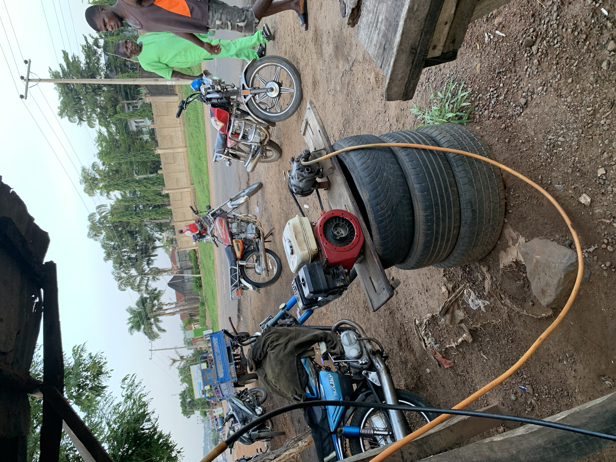 The image shows an outdoor scene in Gbeleko, Nigeria, with several motorcycles and motorcycle-related objects. Three motorcycles are clearly visible, with two others partially in view. Three black tires are stacked on the ground, next to a portable gasoline engine mounted on a wooden platform. An orange hose runs across the ground near the tires. Two men stand near one of the motorcycles; one wears a green outfit, and the other a brown tank top with an orange patch. The ground is dirt and gravel. In the background, trees, buildings, and power lines are visible, suggesting a street setting.