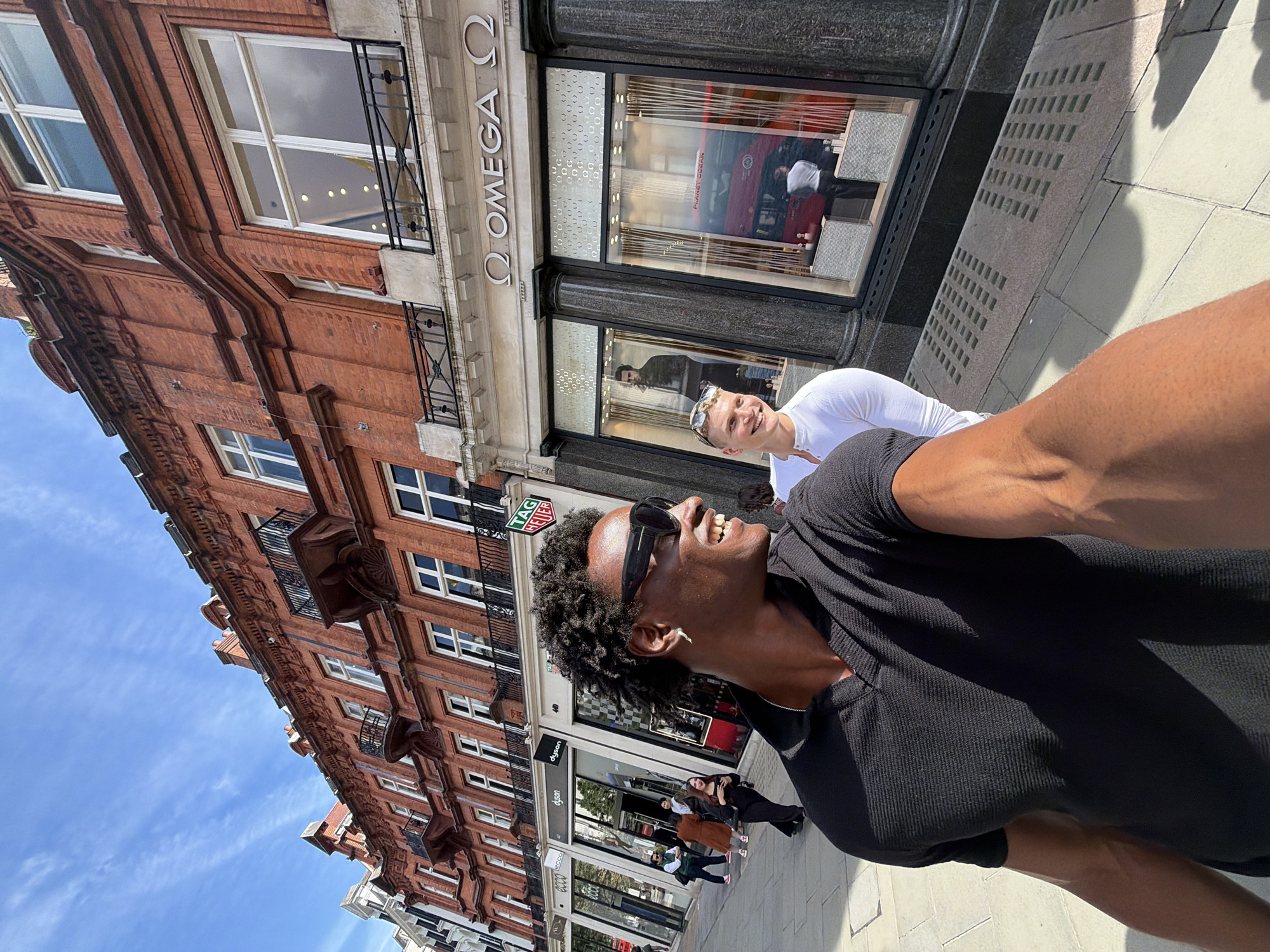 A sunny street scene in London, United Kingdom features two people near an Omega store. The foreground shows a person with dark skin wearing a black shirt and sunglasses, looking upward and smiling. Another person, fair-skinned and wearing a white shirt, stands slightly behind. In the background, a brick building with multiple windows is visible, as is the Omega store with the word "OMEGA" prominently displayed above its entrance. Pedestrians walk along the sidewalk and the sky is blue with some clouds.