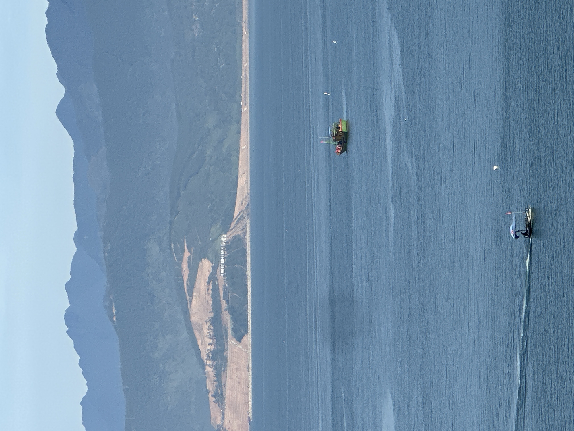 This image shows a wide view of the sea near Da Nang, Vietnam, with mountains in the background. In the middle distance, a green fishing boat is on the water. In the foreground, a smaller boat with a canopy is moving from left to right, leaving a wake. The sky is a light blue. The mountains are layered, with the furthest ones appearing a hazy blue and the closer ones a darker green, with some areas of exposed earth on the slopes. The sea is a deep blue with visible ripples.