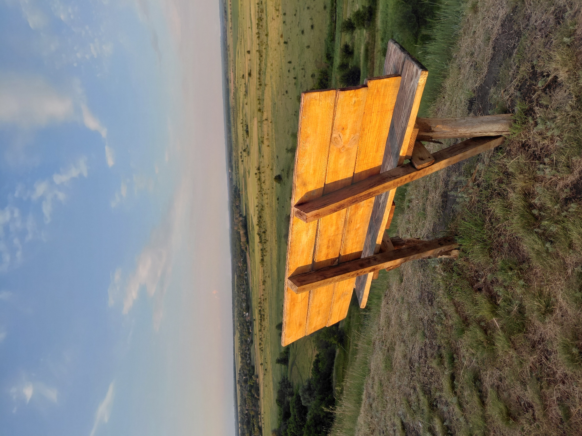 An outdoor scene features a wooden bench on a grassy knoll. The bench is constructed from rough-hewn wooden planks and appears weathered. It is positioned overlooking a vast, rolling landscape that stretches into the distance. The landscape is a patchwork of green fields and darker patches of trees. In the far distance, a faint line of what might be a village or town is visible.
The sky above is a clear blue with scattered white clouds, suggesting fair weather. The lighting indicates it is likely late afternoon or early evening, as the sun is casting long shadows and highlighting the texture of the wooden bench with a warm, golden light. The grass around the bench is a mix of green and dry, brown hues, typical of a natural, untended environment.
There are no people visible in the image, nor is there any text. The overall mood conveyed is one of tranquility and natural beauty, inviting contemplation or rest. The perspective from the bench suggests a viewpoint offering a panoramic vista.