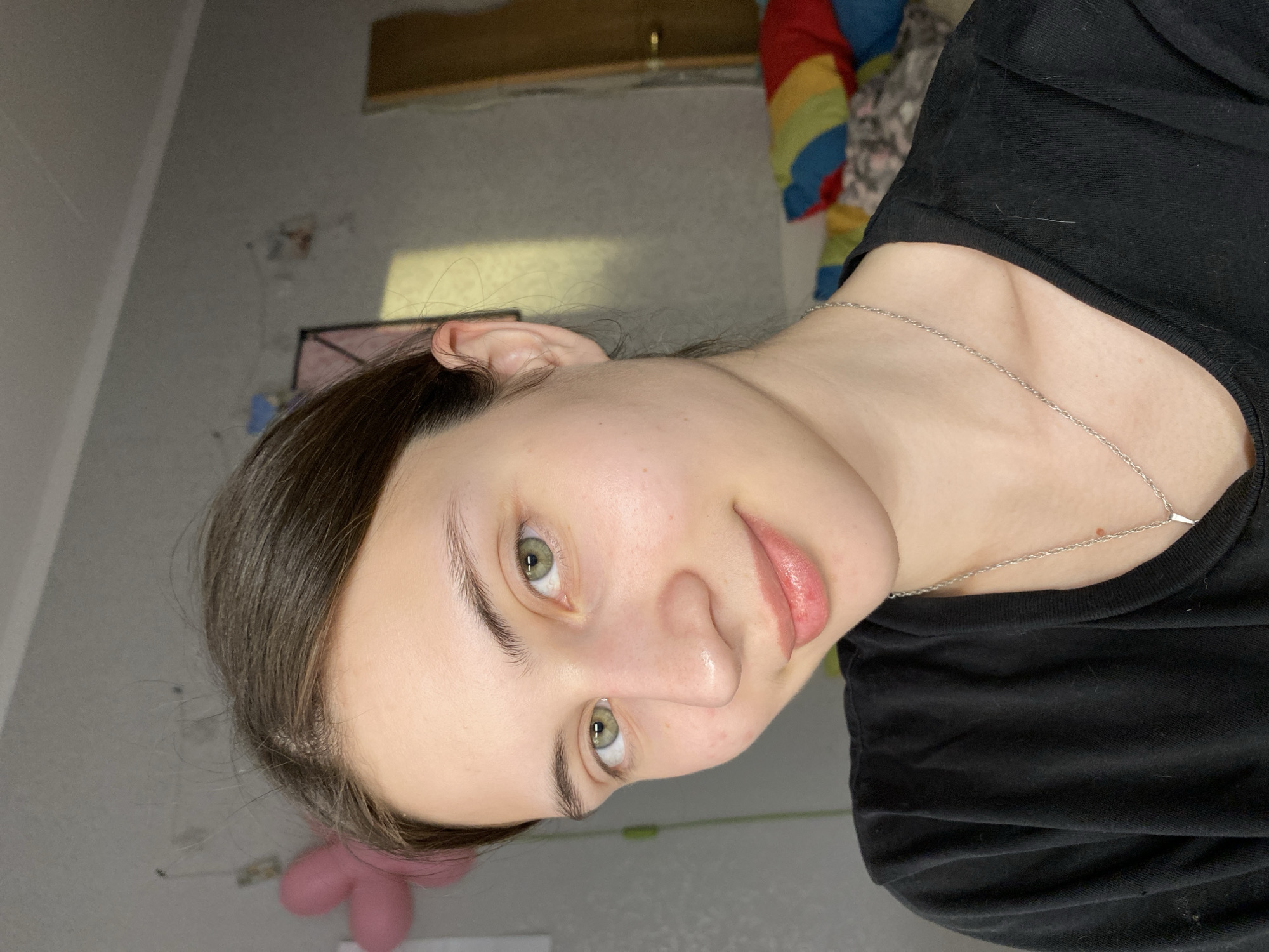 A person with light skin, green eyes, and brown hair is depicted in a close-up shot. The person is wearing a black shirt and a thin silver necklace. The background includes a light gray wall with some visible markings and a wooden fixture near the top. A colorful striped object is also partially visible behind the person. The scene does not provide enough information to determine the specific location in Votkinsk, Russia.