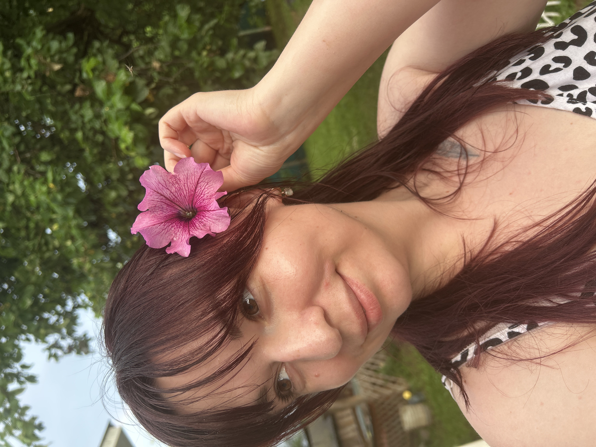 A close-up shot captures a person from the chest up, holding a pink petunia flower to their right ear. The person has dark reddish-brown hair with bangs and is looking to their left. Their skin appears fair with some light freckles. They are wearing a black and white patterned top. The background consists of out-of-focus green foliage and a cloudy sky. The setting appears to be an outdoor, natural environment, possibly a backyard or garden. There is no information to suggest this image is located in Фоки, Russia.