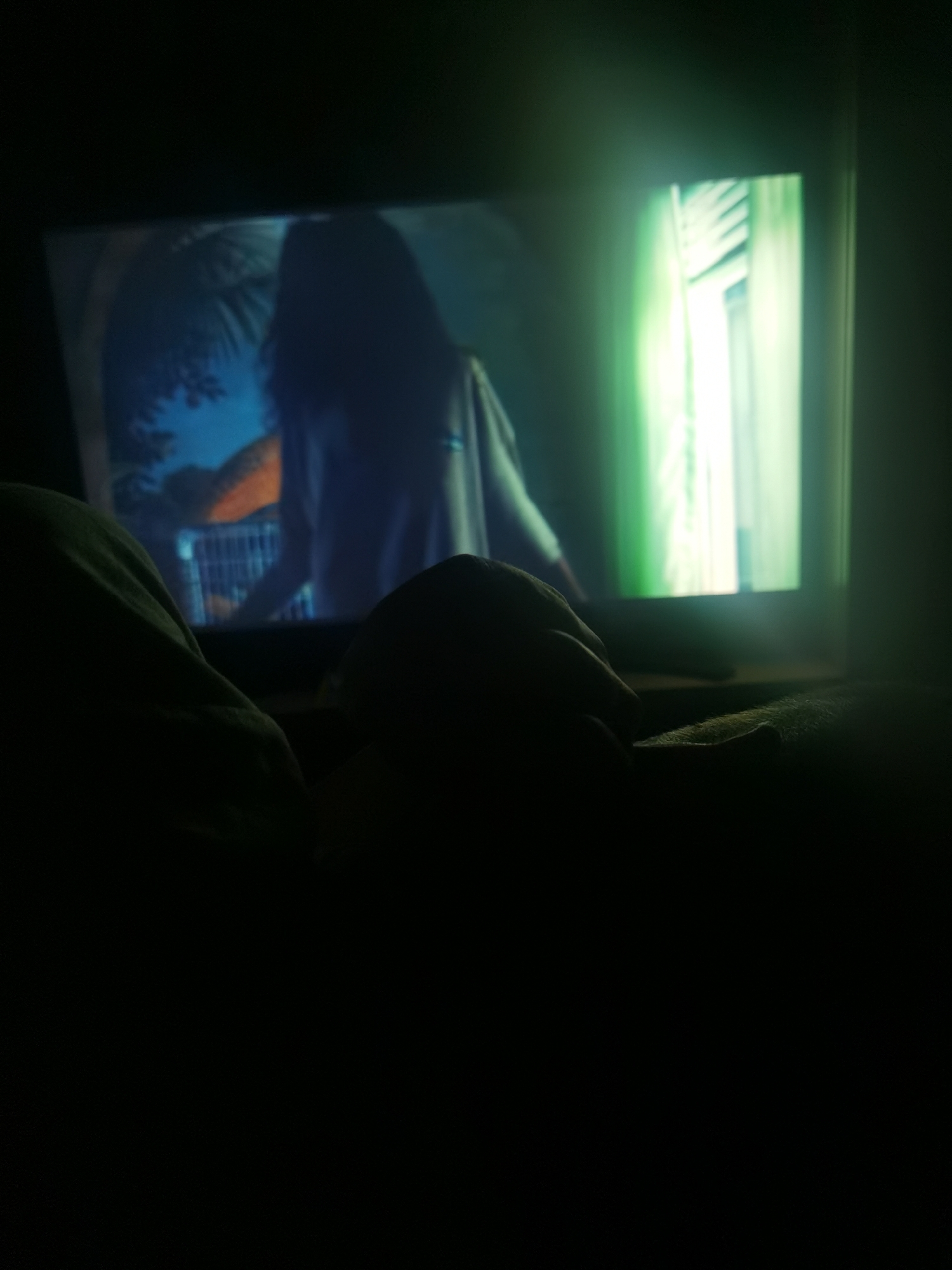 A person is sitting in a dark room, watching television. The television screen shows a woman with long hair, standing in front of a window with white blinds. The setting of the image is unknown, including the possibility that the image was taken in Chaikovsky, Russia.