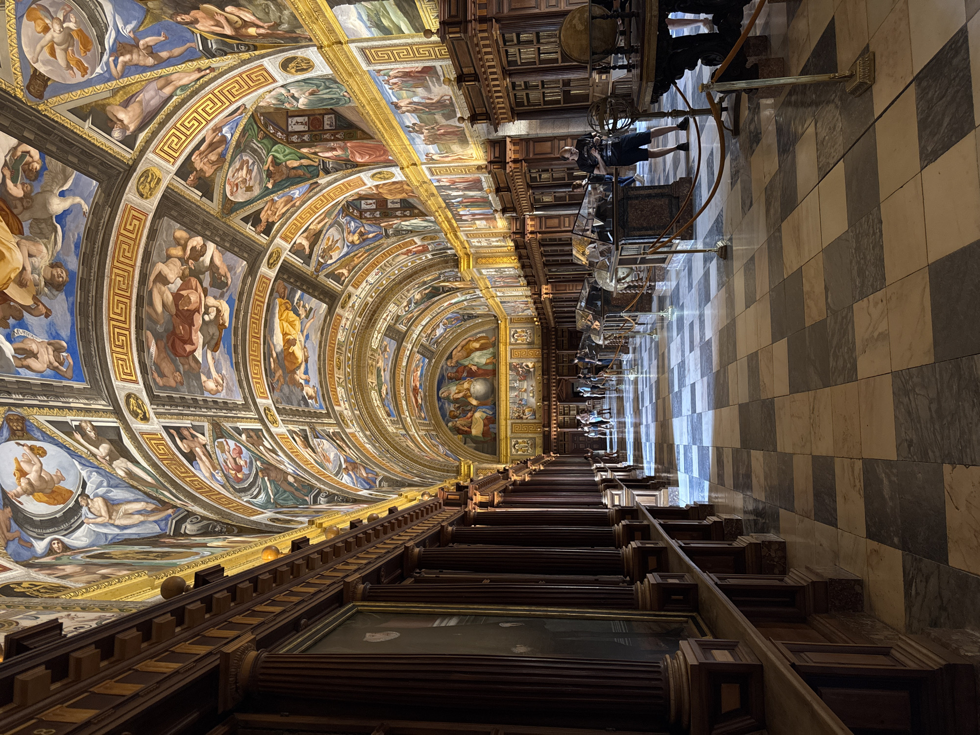 The image depicts the interior of the Royal Library of San Lorenzo de El Escorial, Spain. The vast hall features a grand, barrel-vaulted ceiling adorned with elaborate frescoes depicting mythological and biblical scenes, rendered in vibrant colors and classical style. Along the walls, tall, dark wood bookshelves rise to the ceiling, interspersed with imposing wooden columns and large framed portraits. The floor is paved with a striking checkerboard pattern of light and dark marble tiles, reflecting the light streaming in from unseen windows. Several glass display cases containing globes and other scientific instruments are positioned along the central walkway, some with velvet ropes and brass stanchions for crowd control. A few visitors are visible, scattered throughout the hall, some observing the artwork and displays, while others are in conversation. The overall atmosphere is one of historical grandeur and scholarly pursuit, with a focus on the ornate architecture and rich artistic decoration.