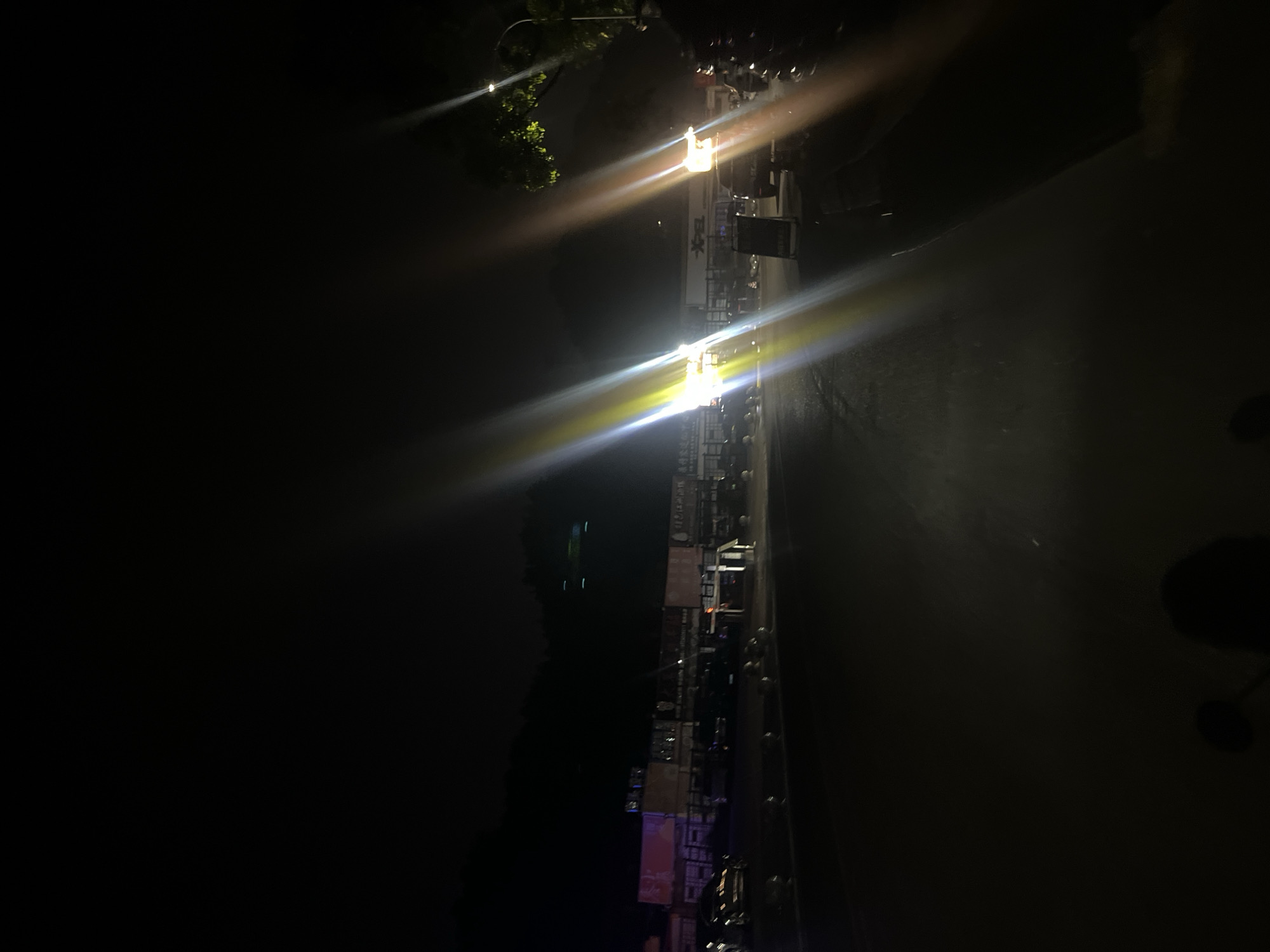The image depicts a nighttime scene in Jiulongpo District, China, characterized by significant artificial illumination and deep shadows. The foreground consists of dark asphalt pavement, possibly a road or parking area, with an indistinct dark shadow resembling a human head and shoulders, potentially from a person operating a vehicle, visible at the bottom center.
The midground features a long, multi-unit commercial or industrial building structure with multiple storefronts or workshops. Several signs with Chinese characters are visible on the building facade; one prominent sign on the right displays the English text "XPEL." Numerous parked vehicles, primarily dark-colored sedans and SUVs, are present in front of the building. Barriers or bollards line the edge of the paved area. Intense, multi-colored lens flares, primarily yellow, white, and orange, originate from bright artificial light sources within the scene, such as vehicle headlights or streetlights, obscuring portions of the view. A dark A-frame sign stands near the "XPEL" building.
The background reveals a dark, mountainous or hilly terrain silhouetted against the night sky, with a few faint, distant lights visible on the slopes. On the far right, a segment of a tree with green foliage is partially illuminated, and a streetlight pole is also visible. No discernable individuals or specific actions are explicitly captured within the scene.