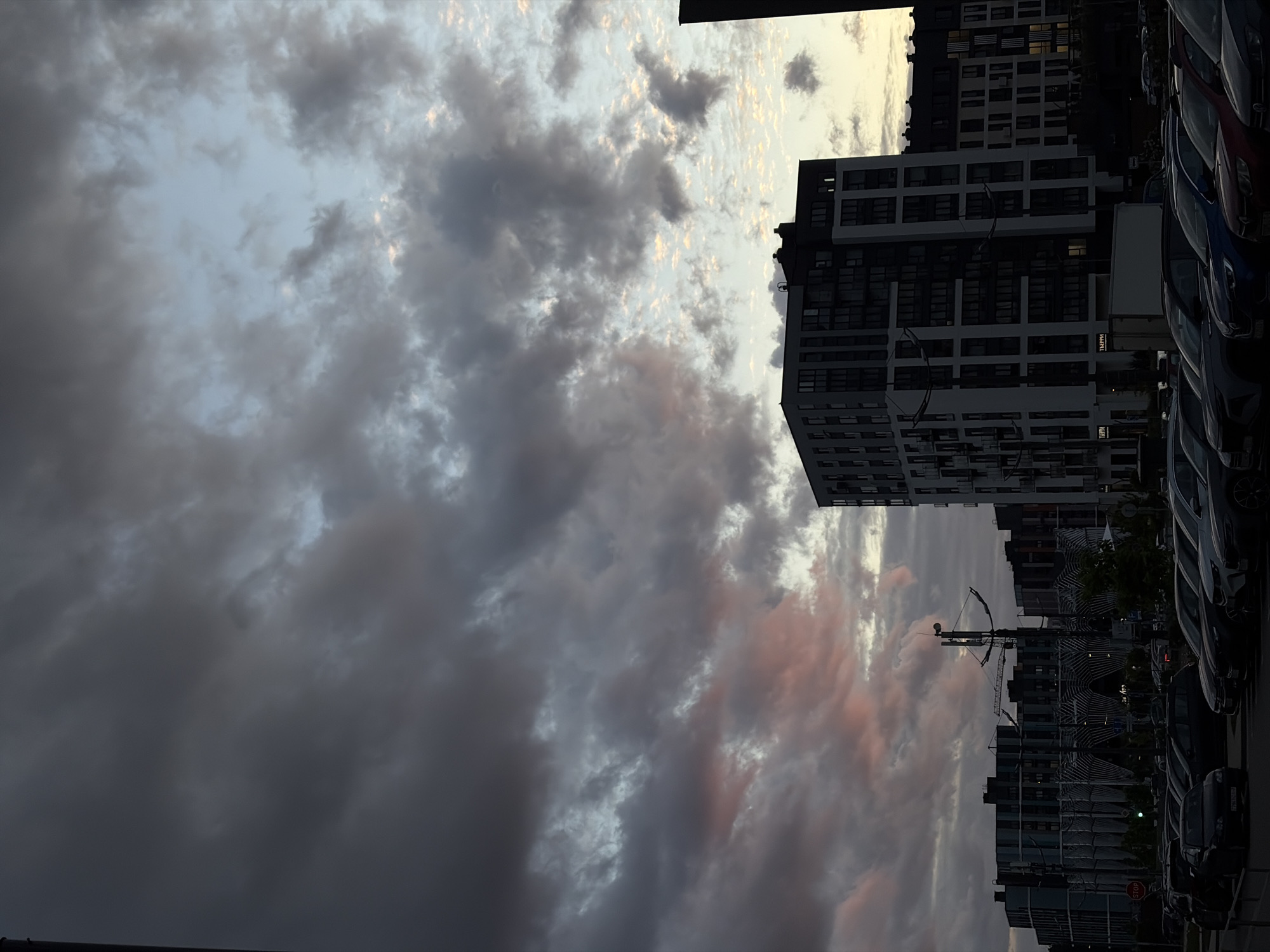 The image shows a cloudy sky above buildings and parked cars in Бараўлянскі сельскі Савет, Belarus. The clouds are dark gray with some lighter patches and pinkish hues near the horizon. Several multi-story buildings are visible, with a modern architectural style. The buildings have many windows and appear to be residential or commercial. A construction crane is visible among the buildings. A row of parked cars lines the right side of the image. The overall scene suggests a late day or early evening atmosphere.