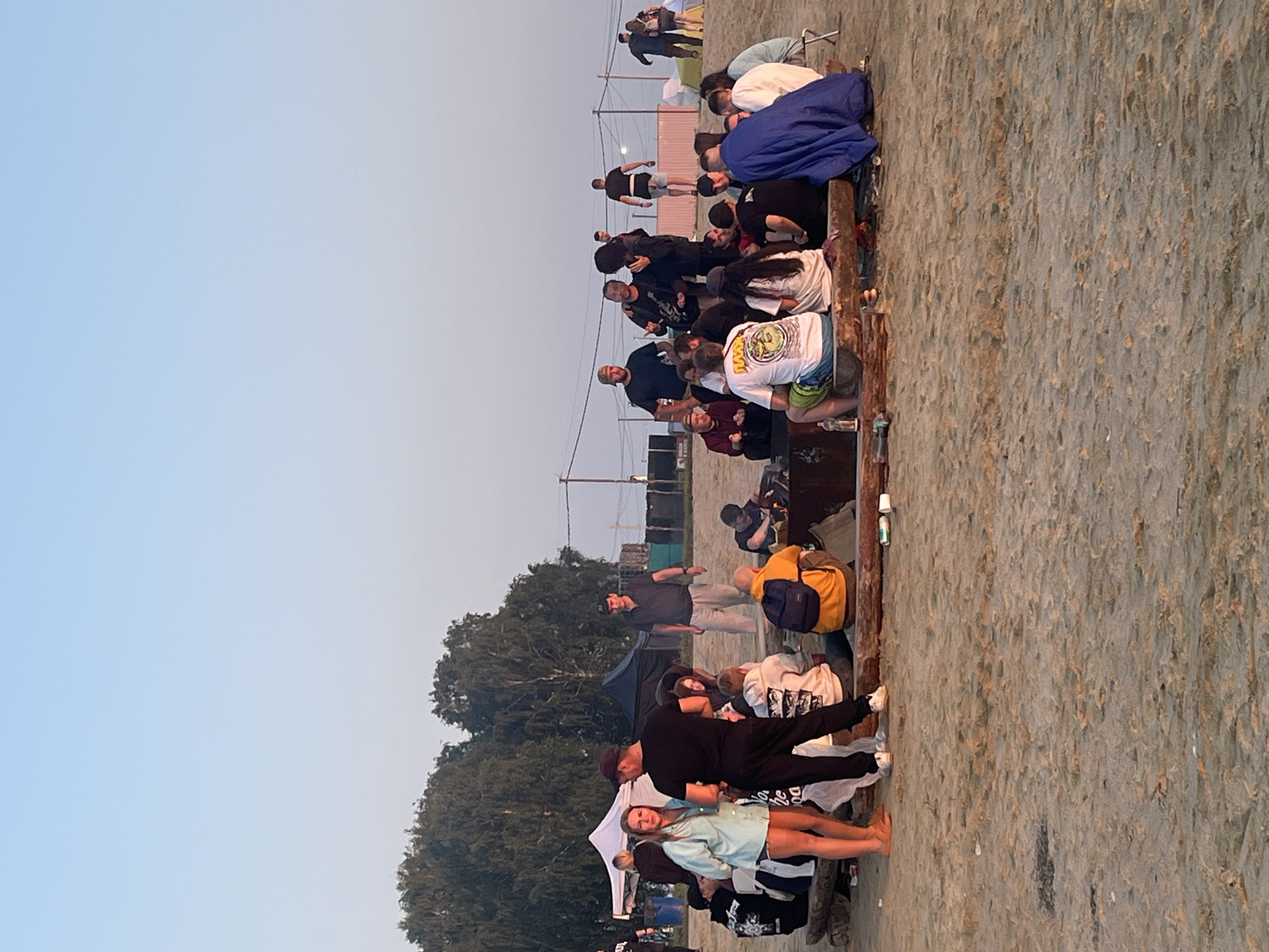 A group of people are gathered around a campfire on a sandy beach in Surakovo, Russia. The sun is setting, casting a warm glow over the scene. In the background, there are trees and tents, suggesting this is a campsite or outdoor event. The people are casually dressed and appear to be enjoying each other's company. Some are sitting on logs around the fire, while others are standing and talking. There are drinks and snacks scattered around the campfire. The overall atmosphere is relaxed and festive.