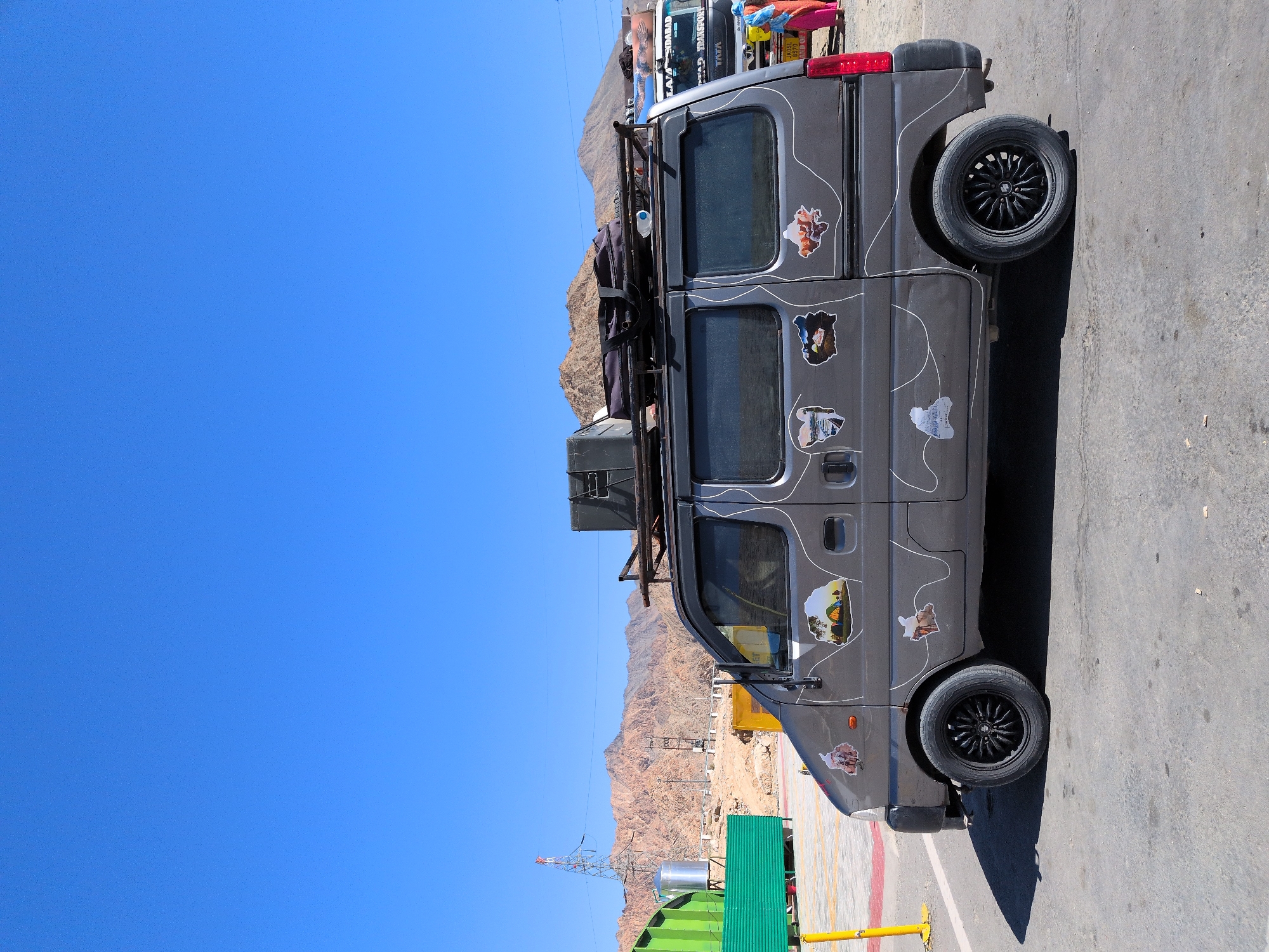 In the city of Chumkar, India, a grey van is parked on an asphalt surface. The van is adorned with various stickers, some depicting landscapes and tents. A luggage rack is mounted on the roof, carrying luggage. In the background, mountains are visible under a clear blue sky. To the left of the van, a green structure and a yellow pole are present. To the right, a truck and two individuals are visible, one wearing a colorful outfit. The scene appears to be a roadside stop in a mountainous region.
