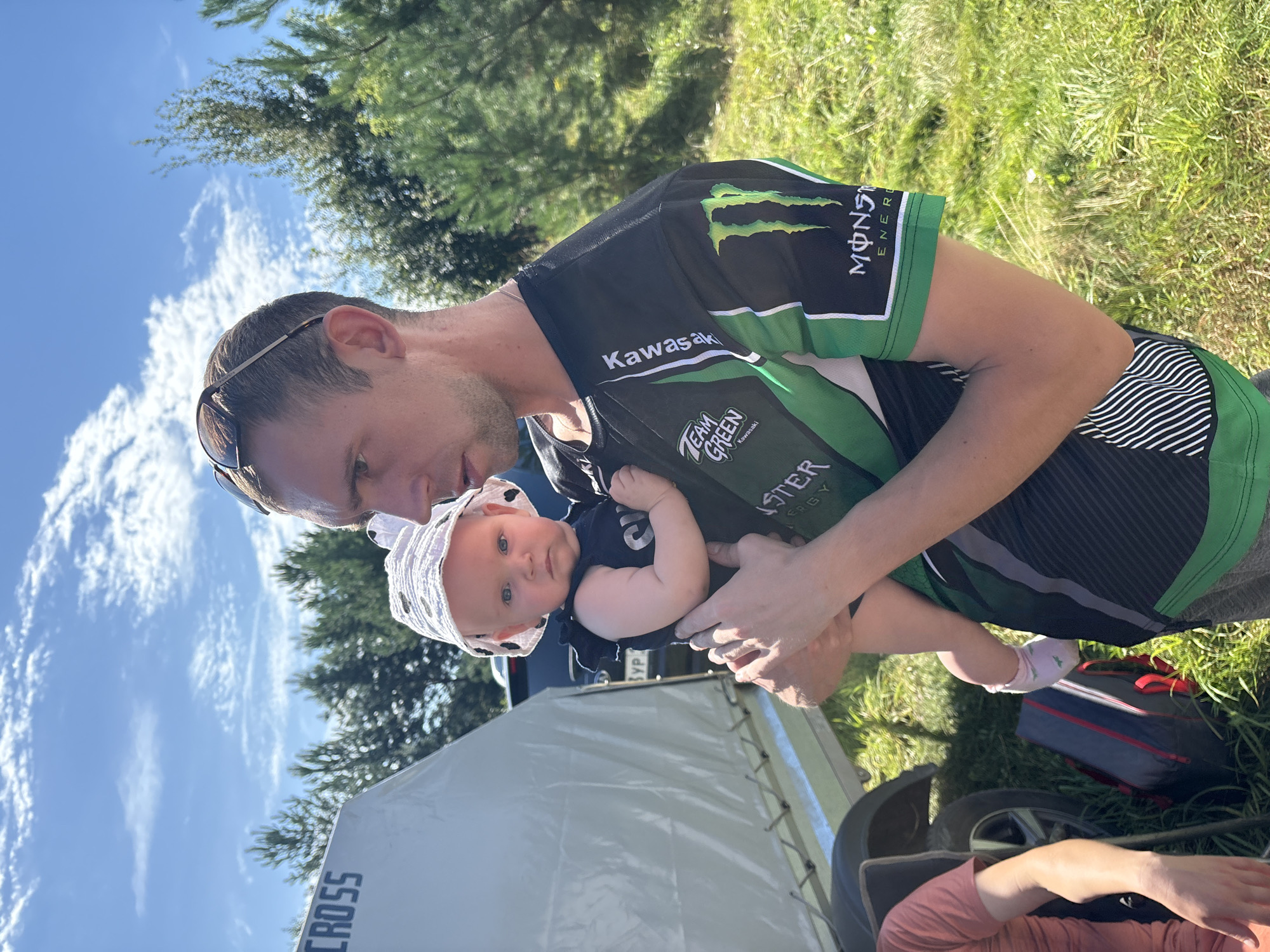 A man holds a baby in an outdoor setting, possibly Votkinsk, Russia, under a bright blue sky with scattered white clouds. The man is wearing a black and green long-sleeved shirt with "Kawasaki" and "Monster Energy" logos. The baby is wearing a white sun hat with black polka dots and a dark-colored outfit. In the background, a white tent or trailer with the word "CROSS" is visible, along with trees and grass. A gray and red bag sits on the ground near a trailer wheel.