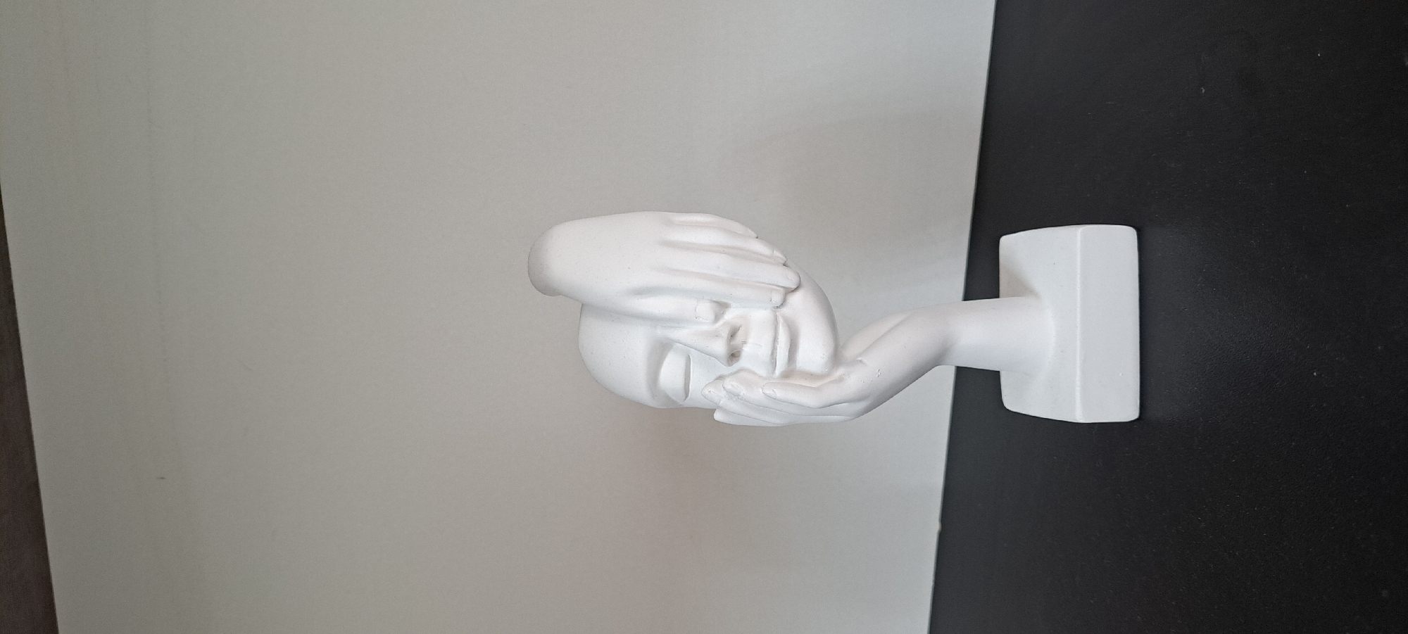 A white plaster bust of a face with closed eyes is positioned on a black surface against a plain, light-colored wall. The bust depicts a face being partially covered by two hands, one covering the eyes and the other resting on the cheek. The bust is mounted on a rectangular base. The scene appears to be indoors, likely on a shelf or table. There is no indication of Yessentuki, Russia, or any specific city location.